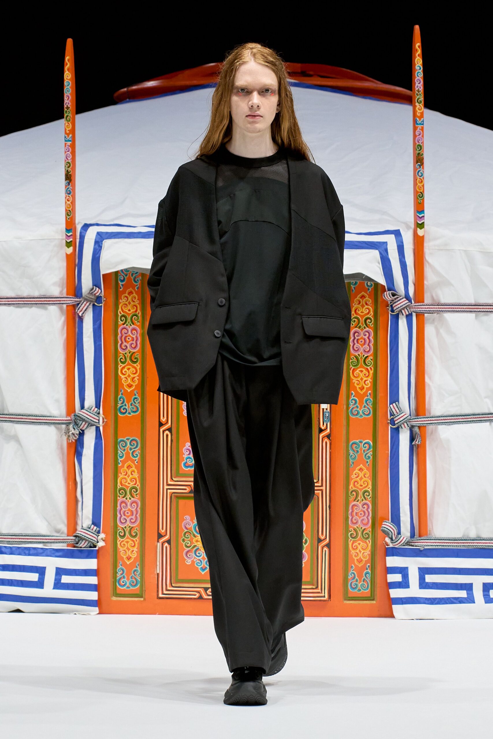 Oversized black jacket pieced in tonal panels with low flap pockets, over a black top with a sheer mesh yoke. Loose black trousers fall in a liquid drape over plain black shoes. Like stepping out before dawn to stoke the stove.