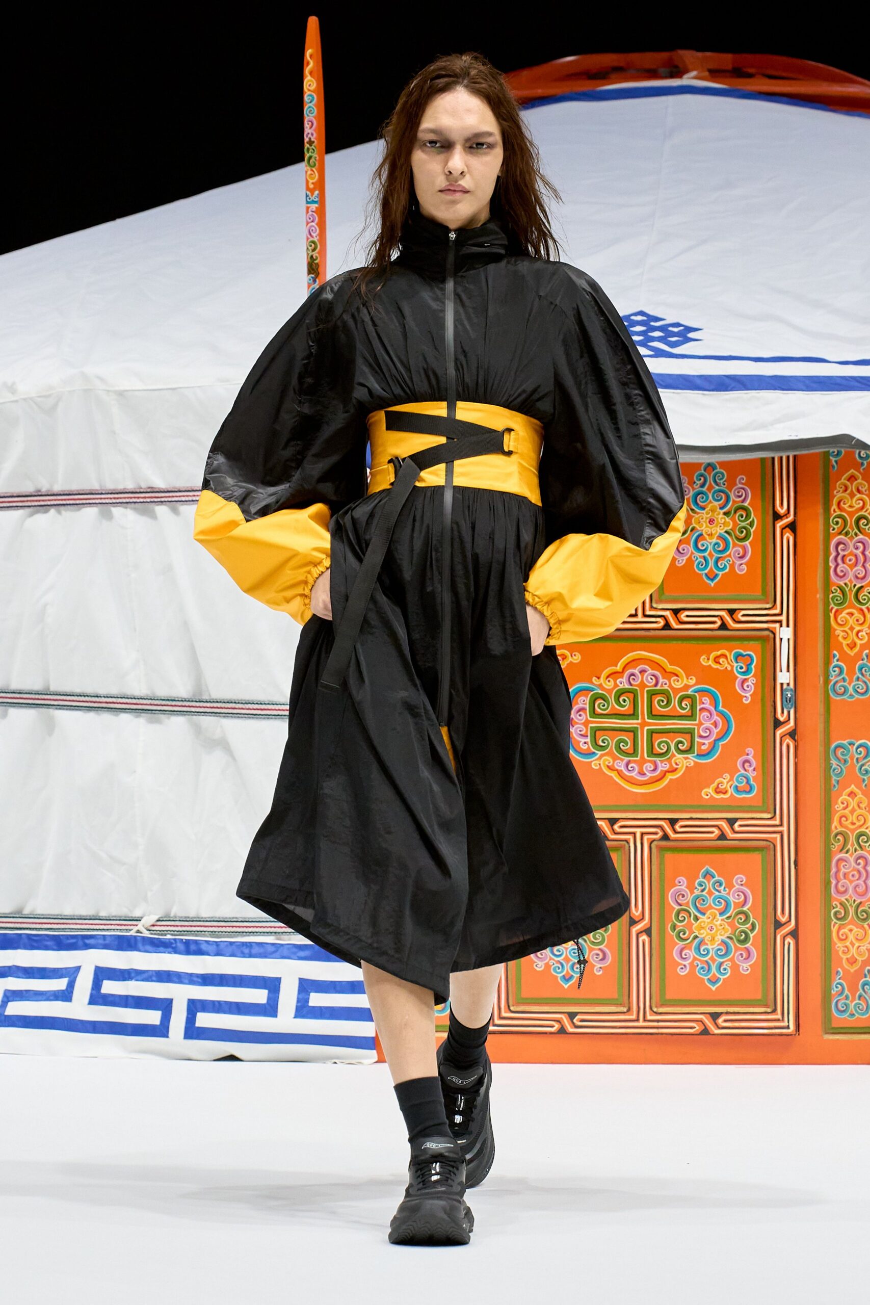 Black ripstop anorak dress with a zip front and gathered volume, its cuffs blocked in marigold. Wide marigold waist cincher with black webbing straps and D-rings. Black trainers, ankle socks. Outside a wind-battered yurt at dusk.