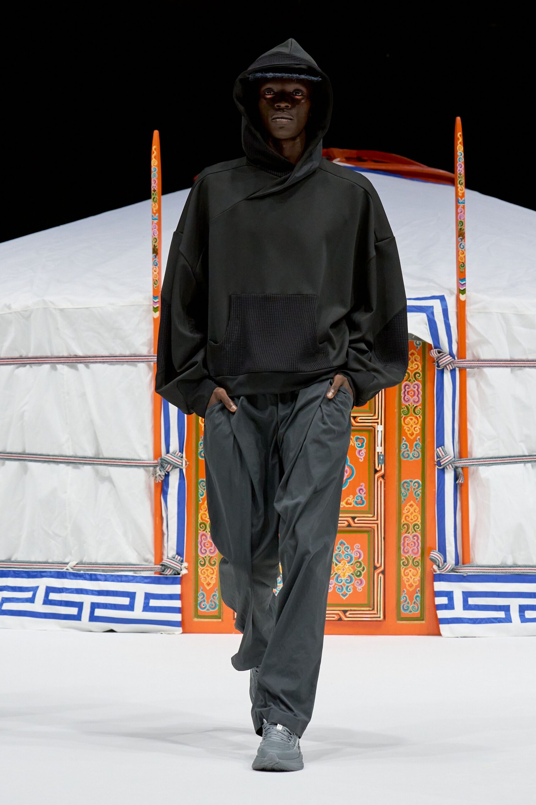 Black hooded pullover in smooth technical cloth, cut with a cross-over neck and waffle-knit pouch pocket. Charcoal wide-leg trousers with deep front pleats, grey sneakers. Like stepping out before dawn to check the camp stove.