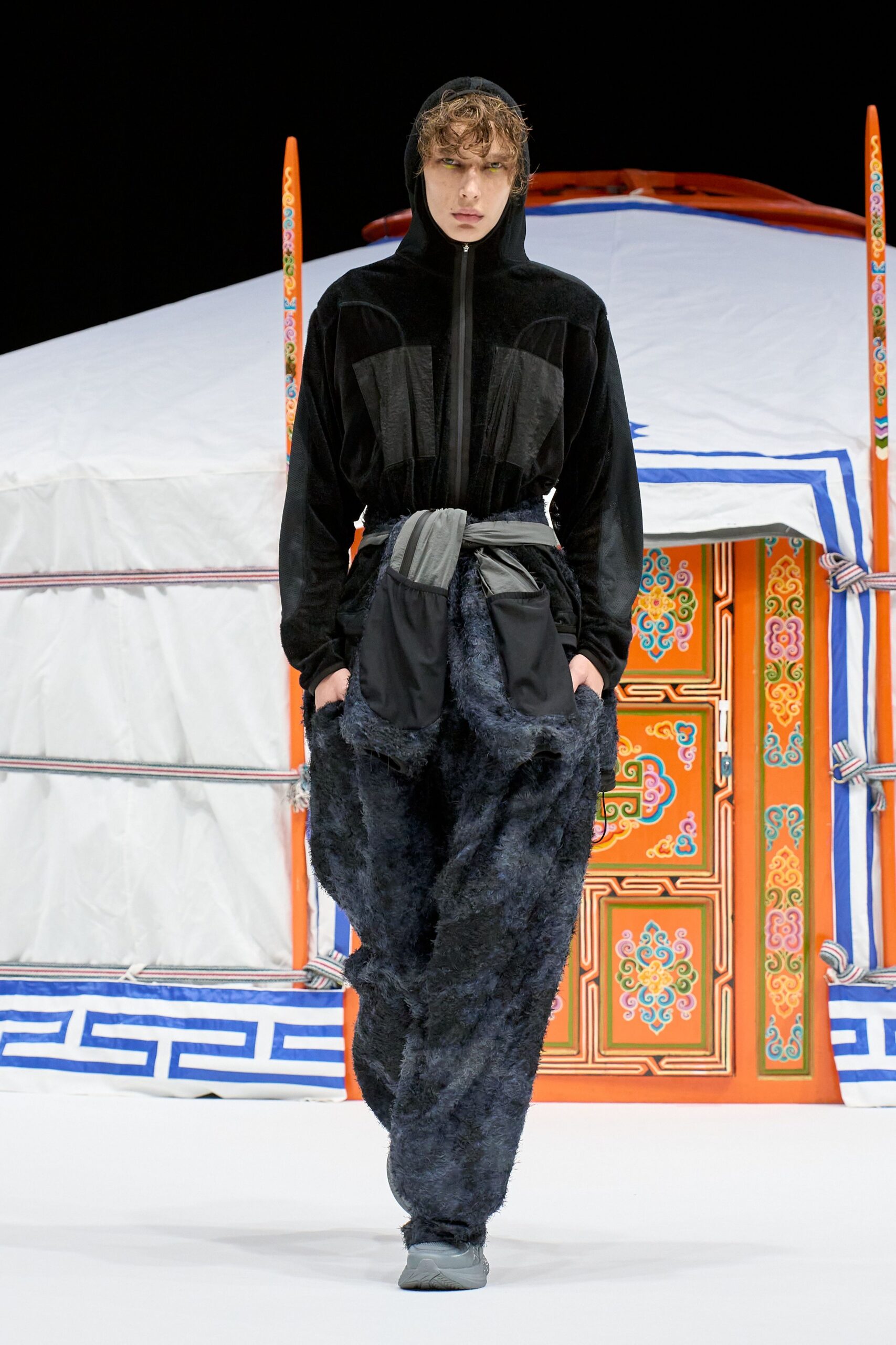 Black velour hooded jacket with a taped front zip and sheer charcoal panels at the chest. Slouchy trousers in shaggy faux fur, tied with a silver ripstop sash and pouch pockets. Dawn outside a winter yurt, kettle just starting.