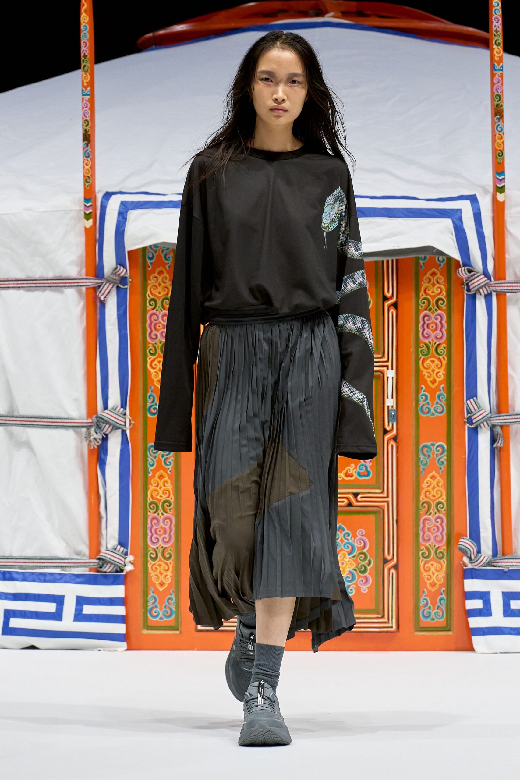 Black long-sleeve jersey top with a iridescent snake curling down one sleeve, the tail dripping at the chest. Charcoal accordion-pleat skirt with a skewed olive panel, ribbed socks and chunky trainers. Crossing a museum courtyard after rain.