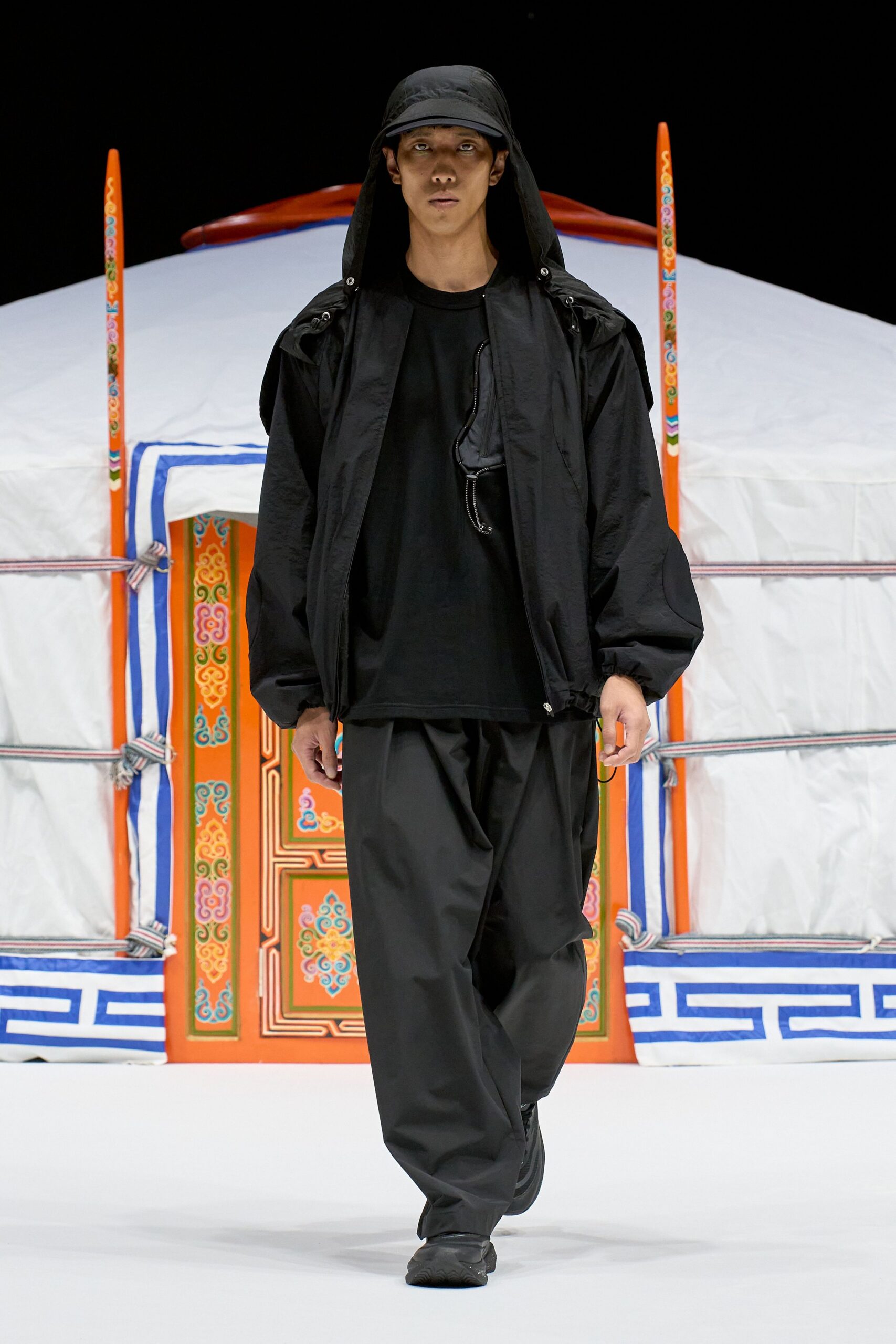 Black nylon hooded jacket with a clipped chest zip detail, over a loose black jersey T-shirt. Wide black technical trousers break heavily at the shoe, with a draped black cap. Stepping out into campsite dark after the stove dies.
