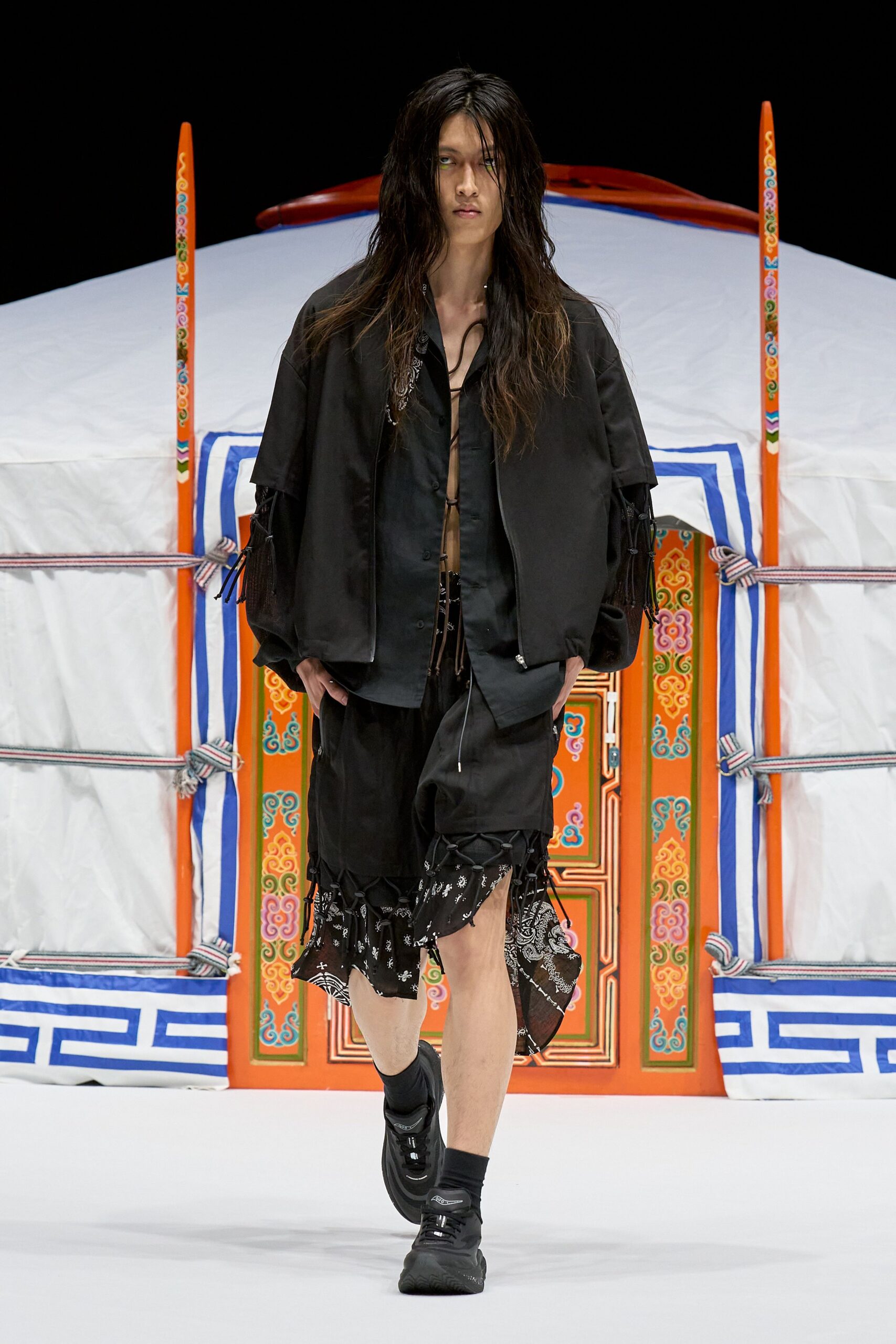 Black cropped jacket with laced mesh sleeves over a black satin shirt left open on cord ties. Black knee-length shorts, bandana-print panels hanging loose at the hem, black sneakers. Outside the yurt at dusk, wind worrying the rope knots.