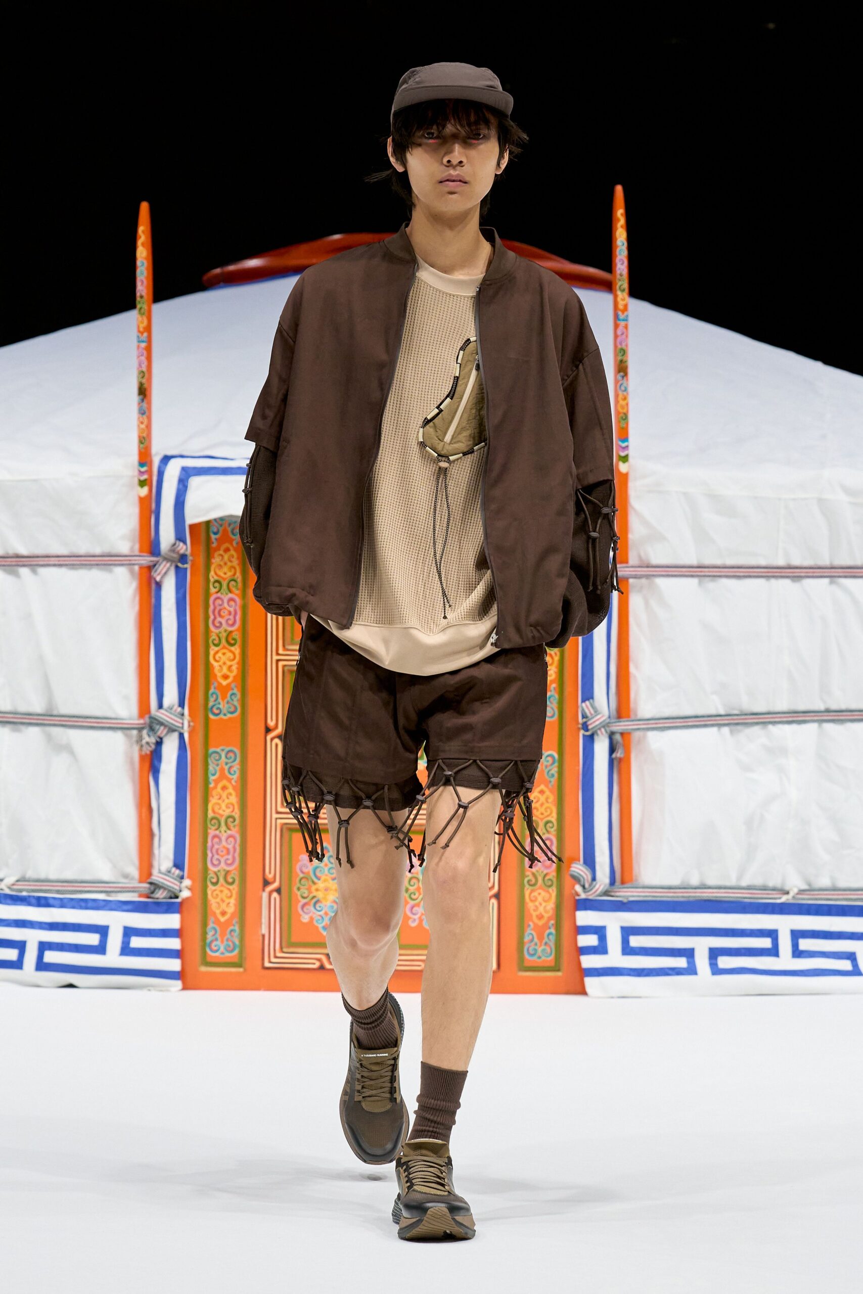 Taupe cap with a flat brim, cocoa bomber with dropped sleeves and cord-laced cutouts, beige waffle-knit tee, small nylon chest pouch, dark shorts fringed with bungee netting, ribbed socks and trail sneakers. Like stepping out of a yurt at dawn.