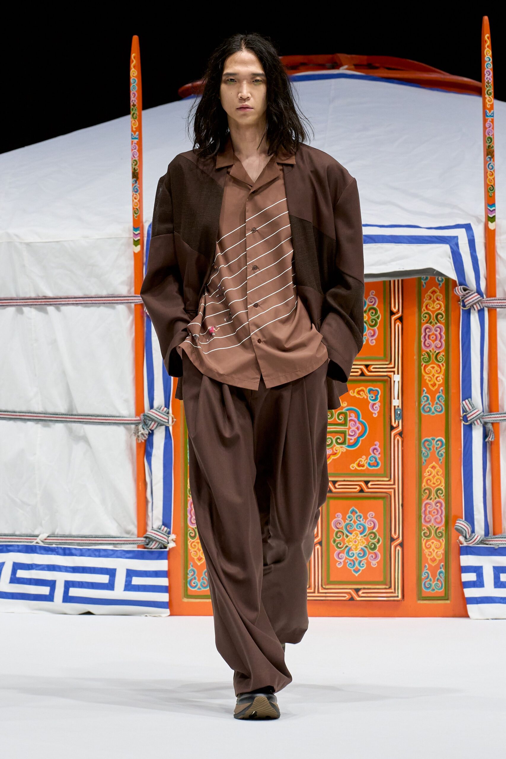 Dark cocoa jacket spliced with open-weave panels over a café-brown camp shirt cut with slanting white pinstripes and a tiny pink embroidered sprig. Fluid wide-leg trousers, black trainers. Crossing a museum atrium before the rain starts.