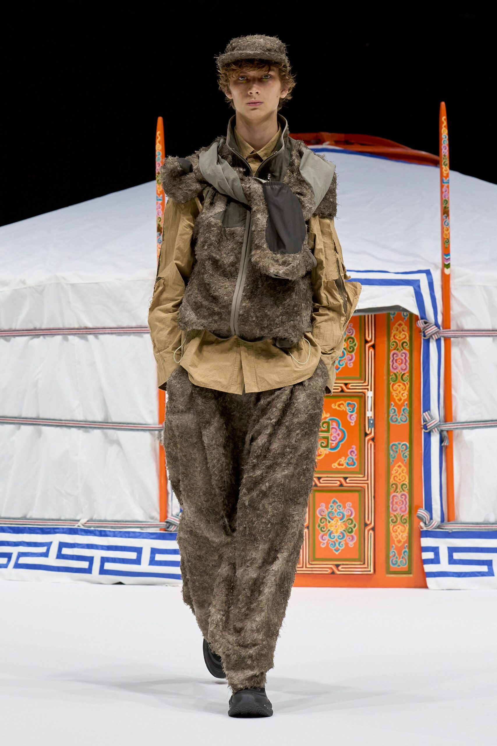 Brown shag-pile cap, sand crinkled nylon shirt, and a taupe faux-fur vest with a rubberized zip and pouchy chest pocket. Matching furry trousers bunch at the ankle over black slip-ons. Dawn outside a yurt, air sharp enough to sting.