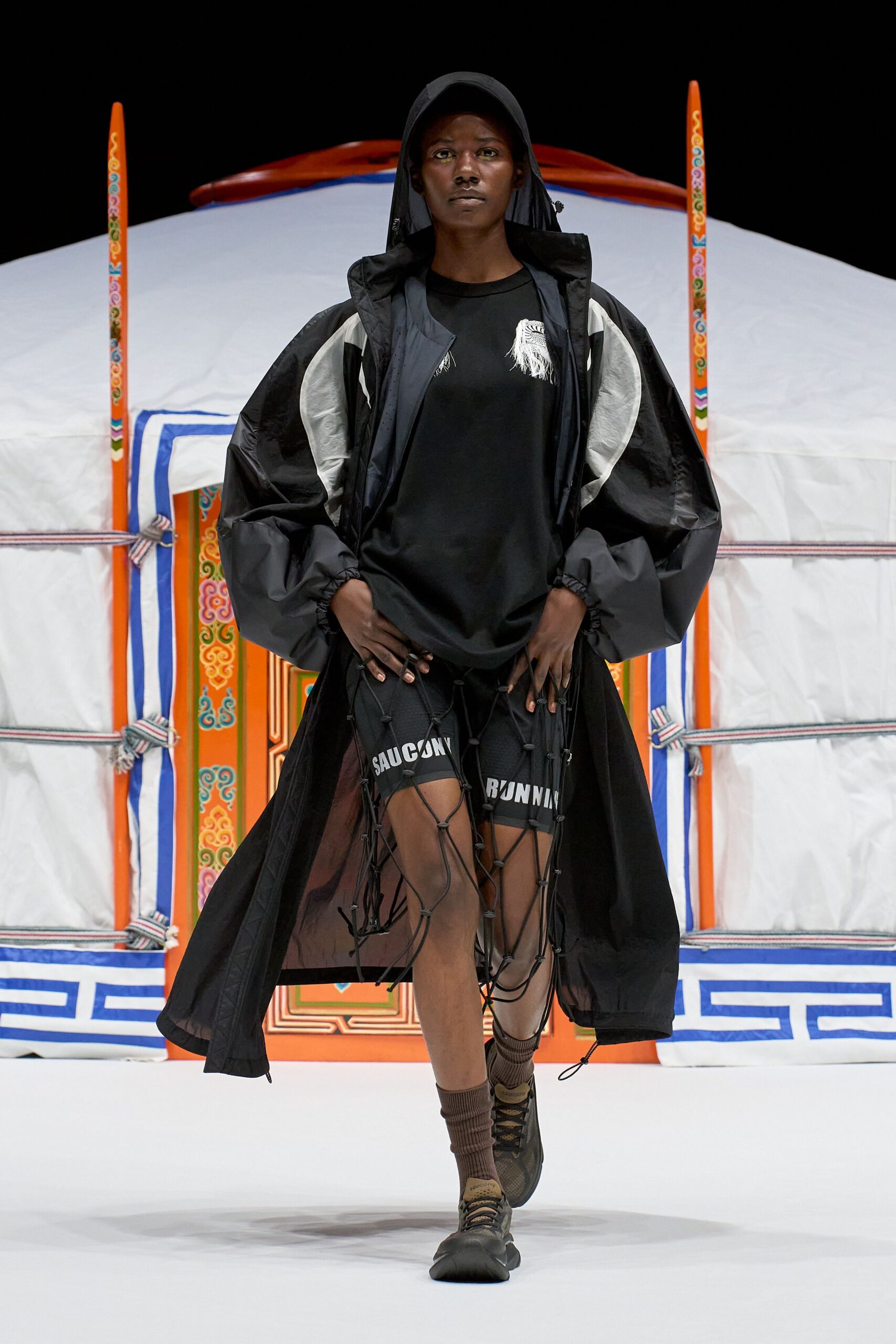 Black nylon hooded parka with silver crescent panels and balloon cuffs, loose black tee with a frayed white chest patch, black running shorts trapped under bungee netting, ribbed socks and trail shoes. Like stepping out of a yurt at dawn.