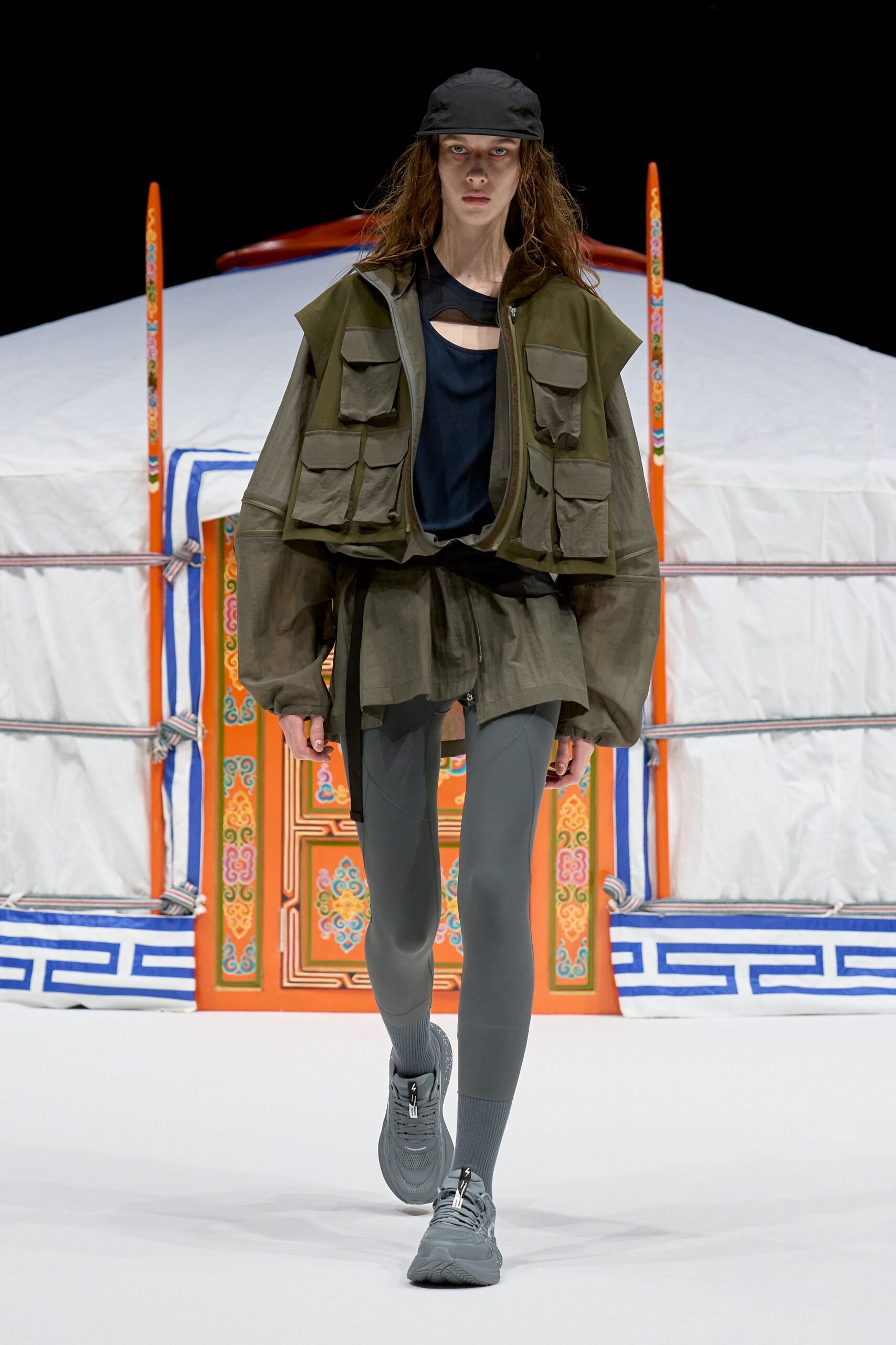 Black nylon cap, olive utility vest with crinkled flap pockets over a black cutout tank, sheer smoke-gray anorak with balloon sleeves, slate leggings, ribbed socks and gray mesh trainers. Like stepping out for dawn tea beside a wind-whipped camp.