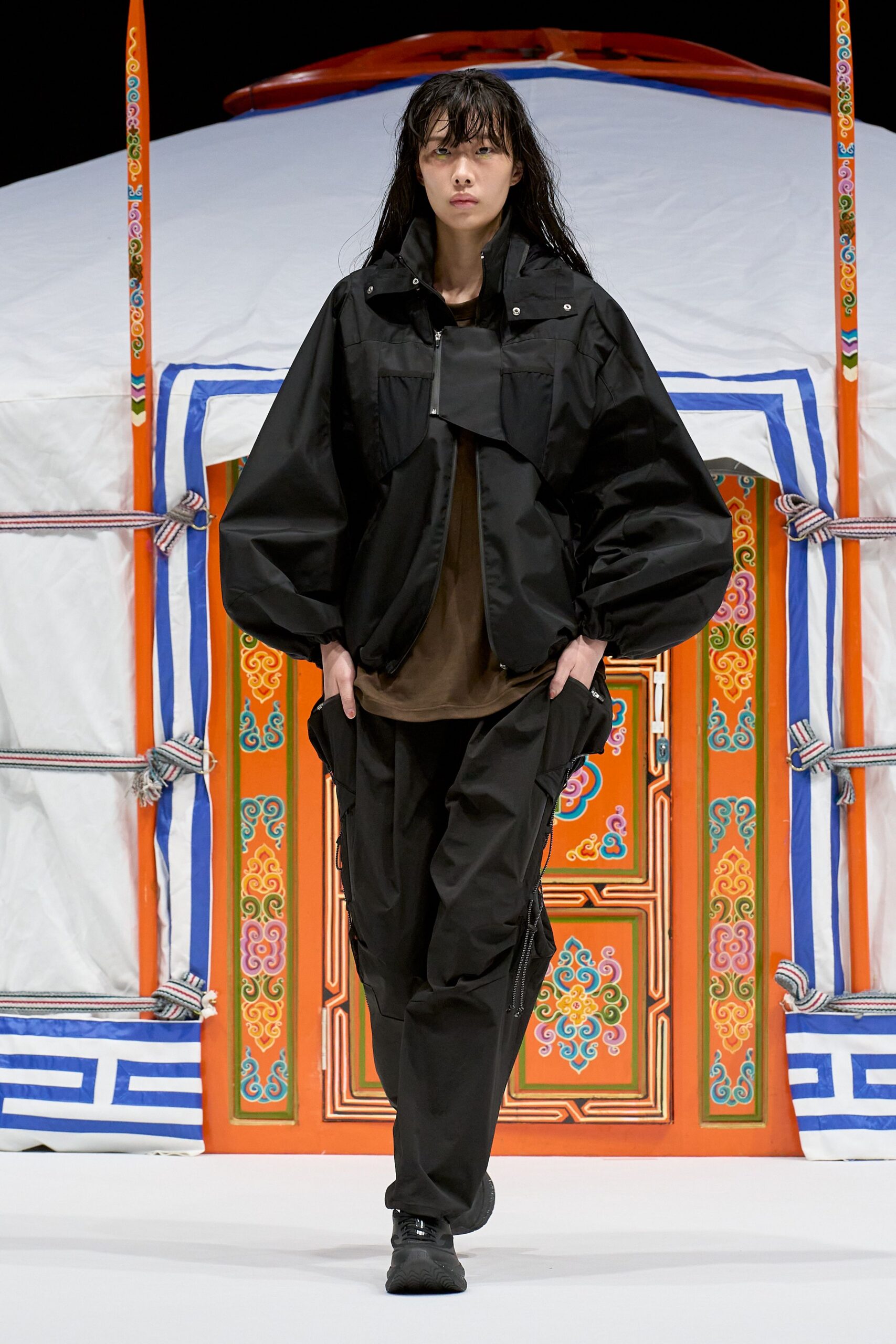 Black technical shell jacket with a snapped storm flap and balloon sleeves, loose umber jersey tee, black cargo trousers traced with long side zips, black trail shoes. Like stepping out of a tent before dawn in cold dust.