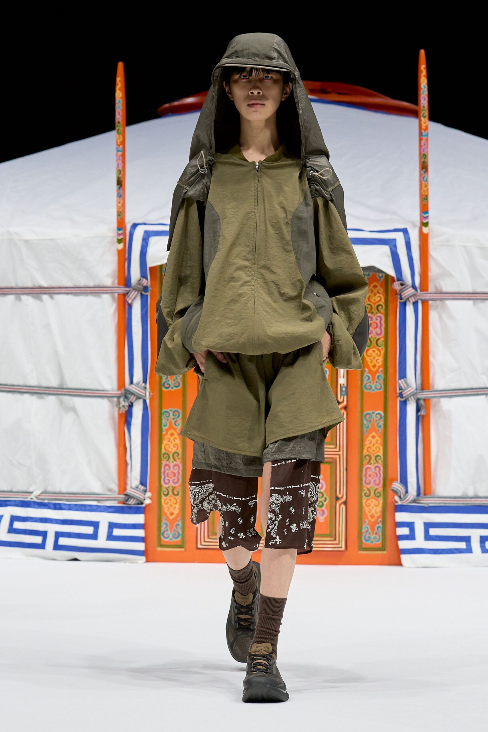 Olive crinkled nylon hooded jacket with a zip front and dangling drawcord loops at the shoulders, layered over split-panel shorts and brown paisley underlayers. Ribbed socks, trail sneakers. Just outside the yurt at first light.