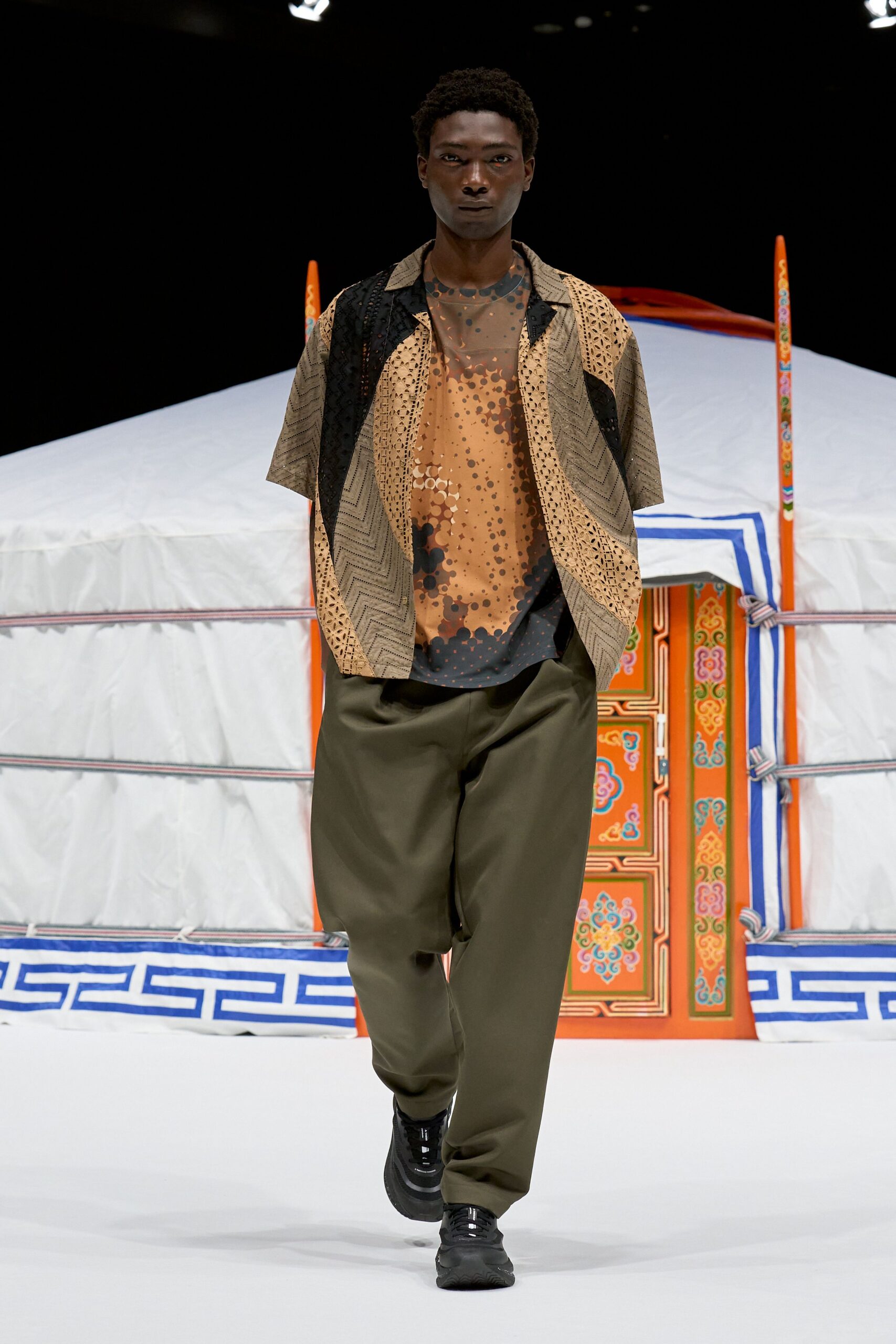 Patchwork camp shirt with eyelet panels and chevron-woven sections over a sheer mesh tee printed with smoke-brown dots. Olive trousers cut wide through the leg, black trainers. Waiting by the station kiosk after rain.