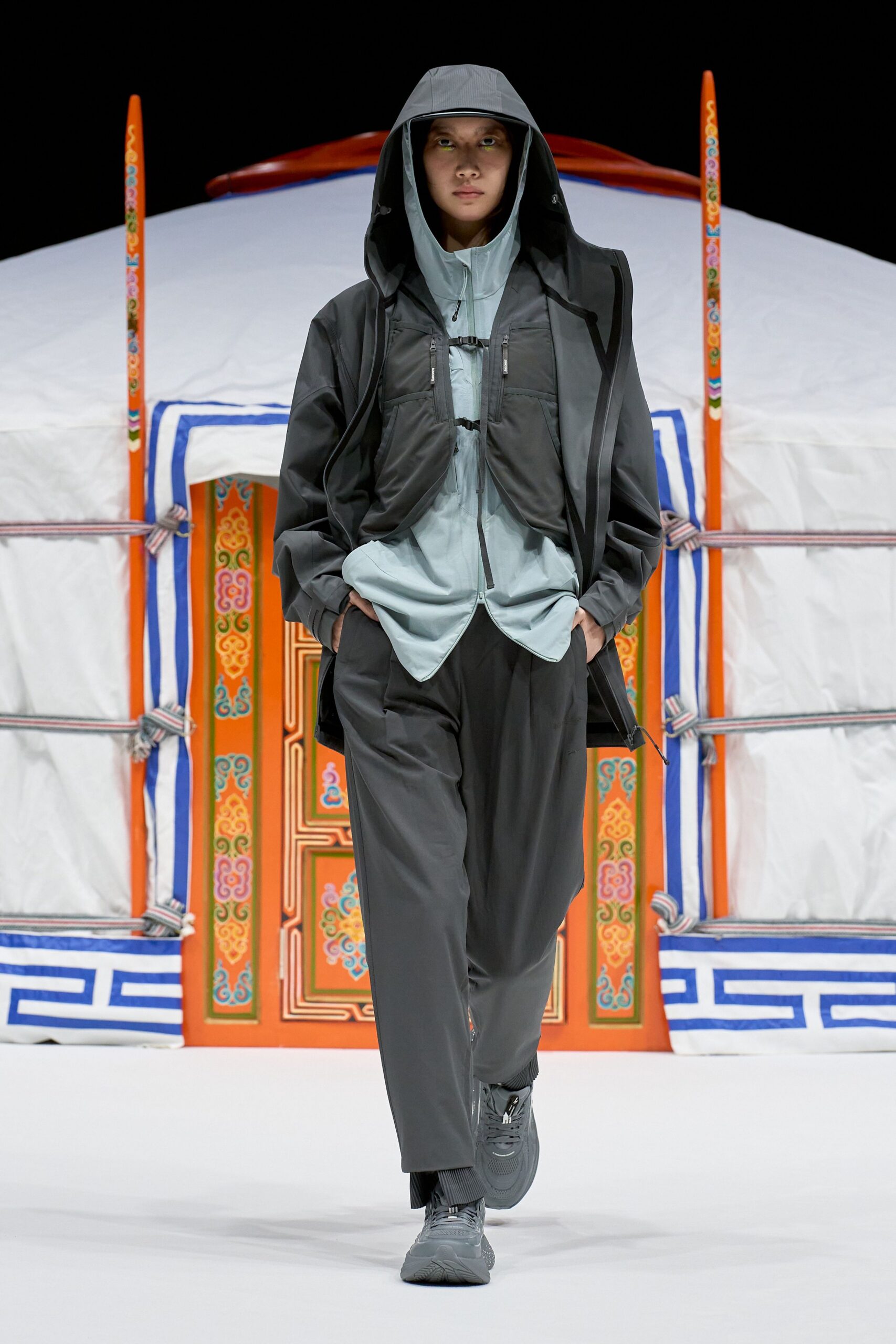 Charcoal hooded shell coat with taped seams over a pale grey zip windbreaker cinched by two tiny buckle straps. Loose charcoal trousers with a pressed crease, ribbed socks, matte trainers. Dawn at a trailhead, map still folded.