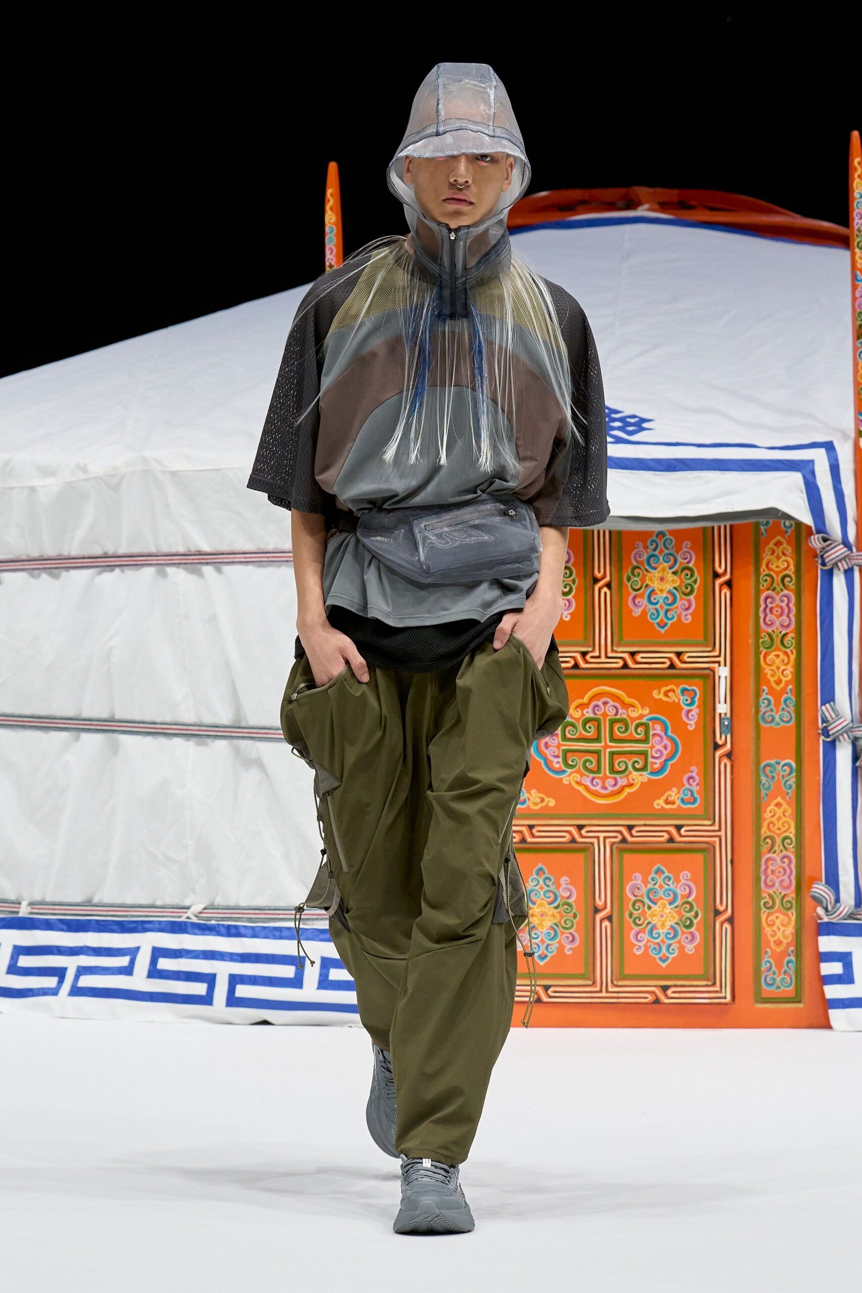 Translucent ripstop bucket hat with a taped seam brim, paneled mesh tee with a high zip neck, smoky PVC belt bag, olive cargo trousers with dangling drawcord hems, grey trainers. Like stepping out of a tent before dawn.