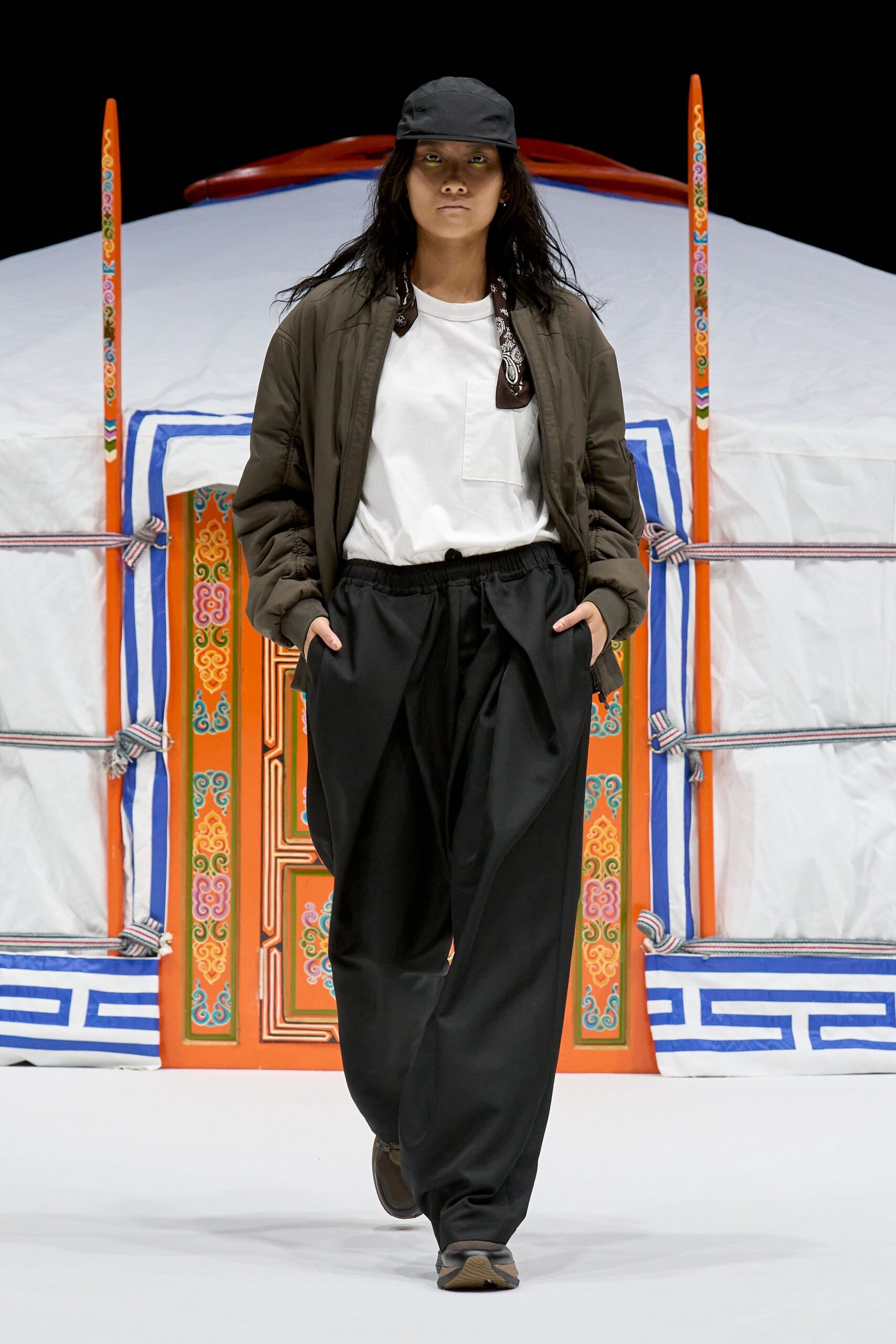 Black nylon cap, olive bomber jacket with ruched sleeves, white crewneck tee with a square patch pocket, dark bandana knotted at the neck, black trousers with a gathered drawstring waist, brown trainers. Dawn outside a roadside yurt.