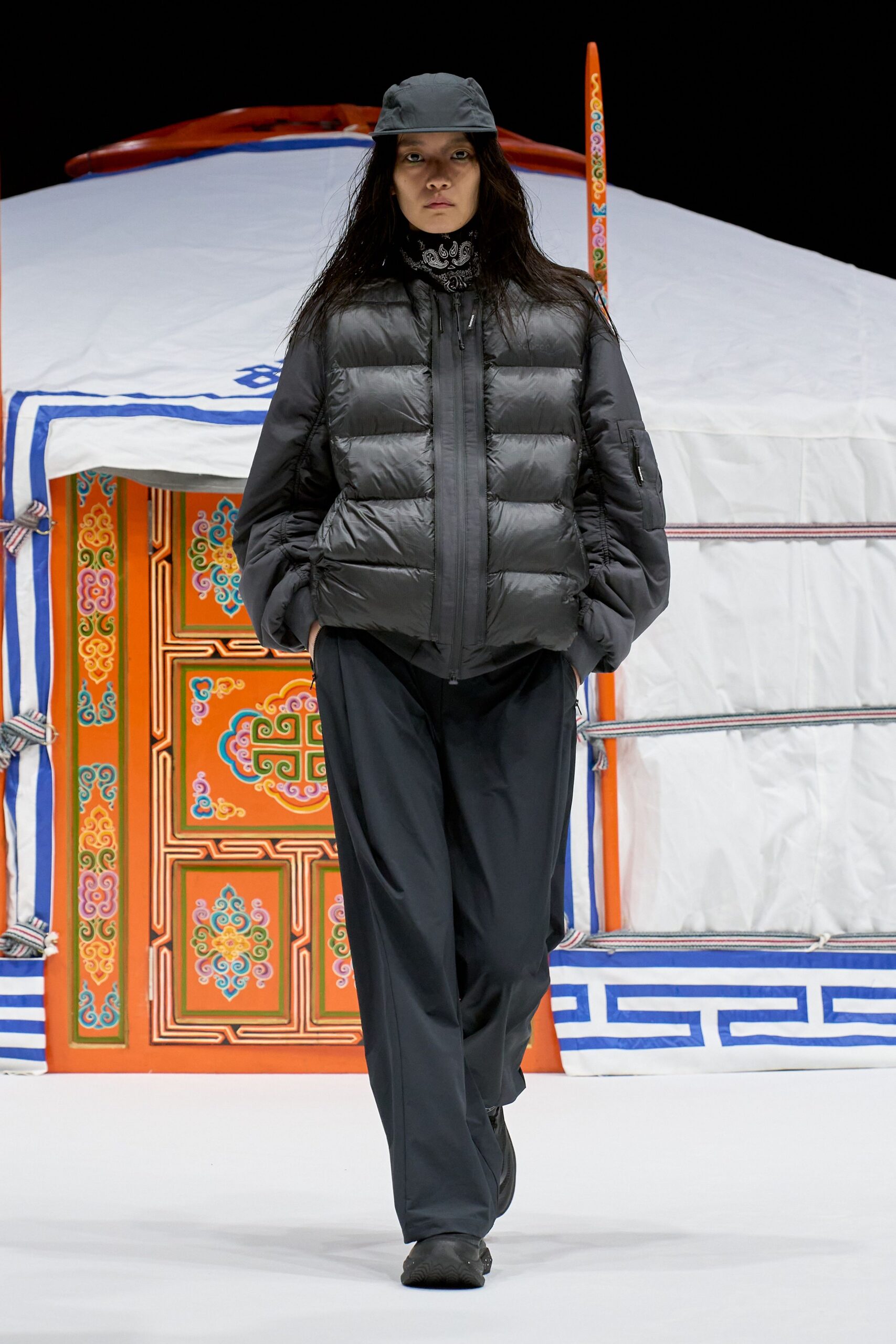 Charcoal nylon cap, black paisley bandana at the neck, matte shell puffer jacket with wide quilted baffles, fluid black trousers breaking over rubber-soled shoes. Like stepping out before dawn to check the stove in a winter camp.