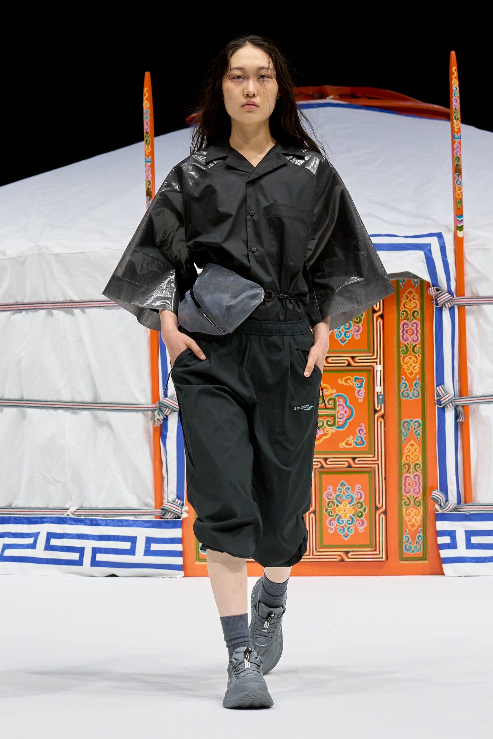 Black camp-collar shirt with glossy taped shoulder panels and wide cropped sleeves, cinched at the waist. Black cropped track pants with an elastic waist, slate pouch bag, charcoal trainers. Crossing wet tarmac between flights.