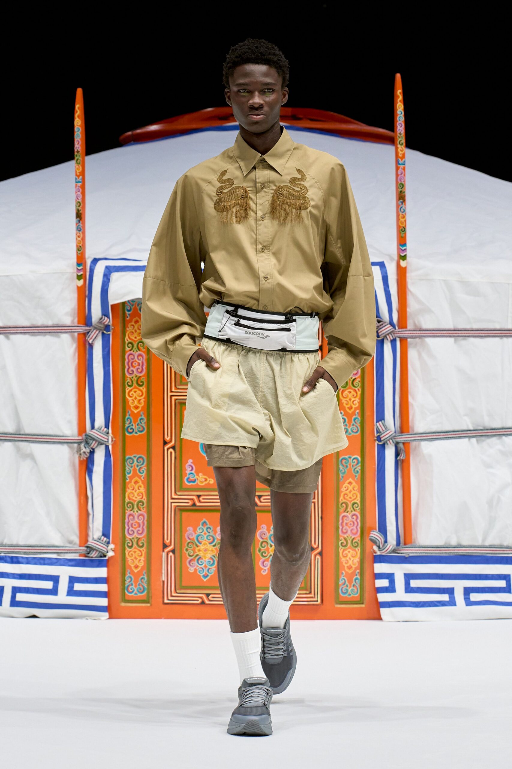 Khaki poplin shirt with snake embroidery and fringe across the chest, pale nylon running shorts over taupe under-shorts, white zip belt bag, ribbed tube socks, grey mesh trainers. Like dawn laps around a desert camp.