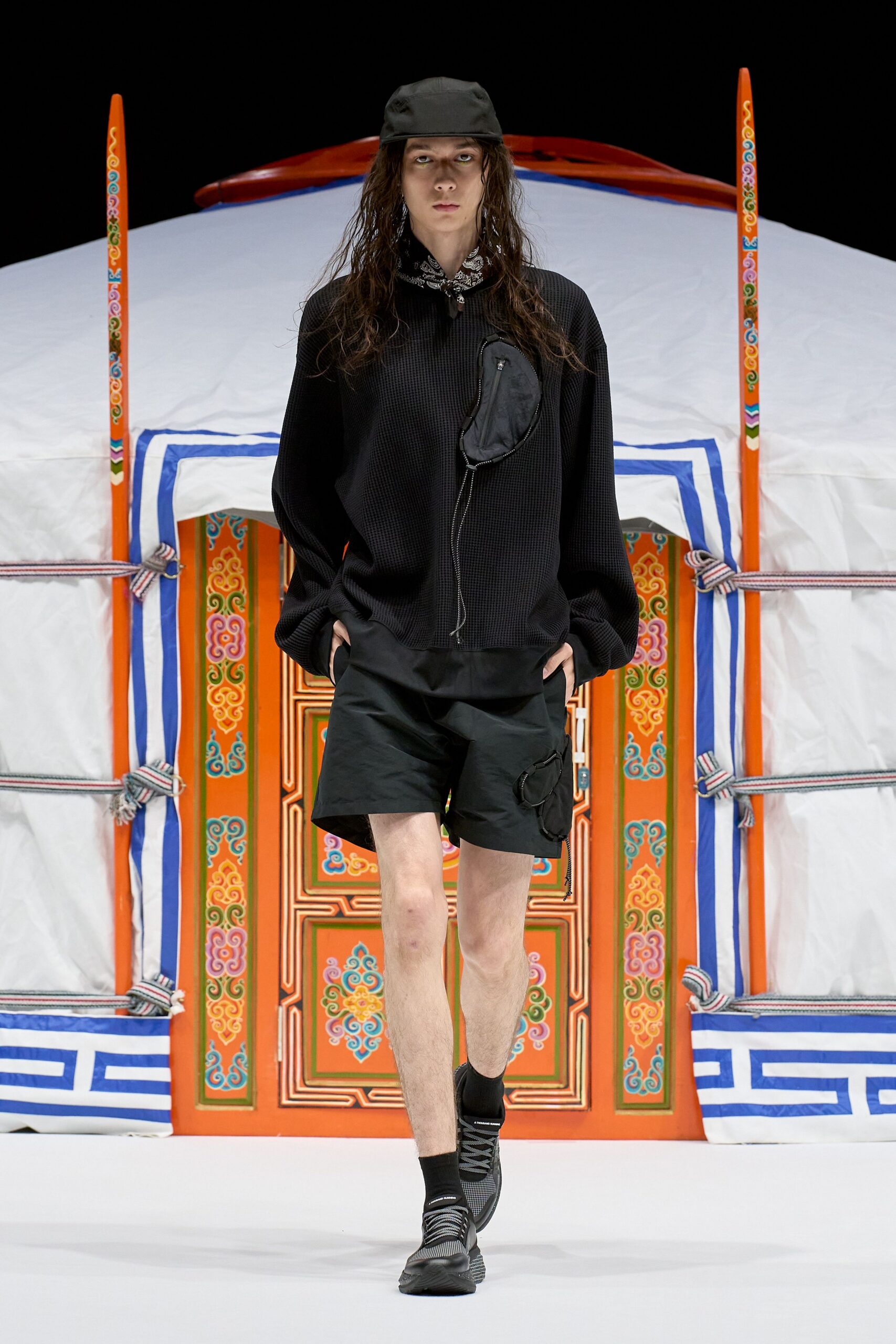 Black nylon cap, paisley bandana knotted at the throat. Black waffle-knit pullover with a zip pouch snaking down the chest. Black nylon shorts with a thigh pocket, mesh trainers. Stepping out for instant noodles after rain.