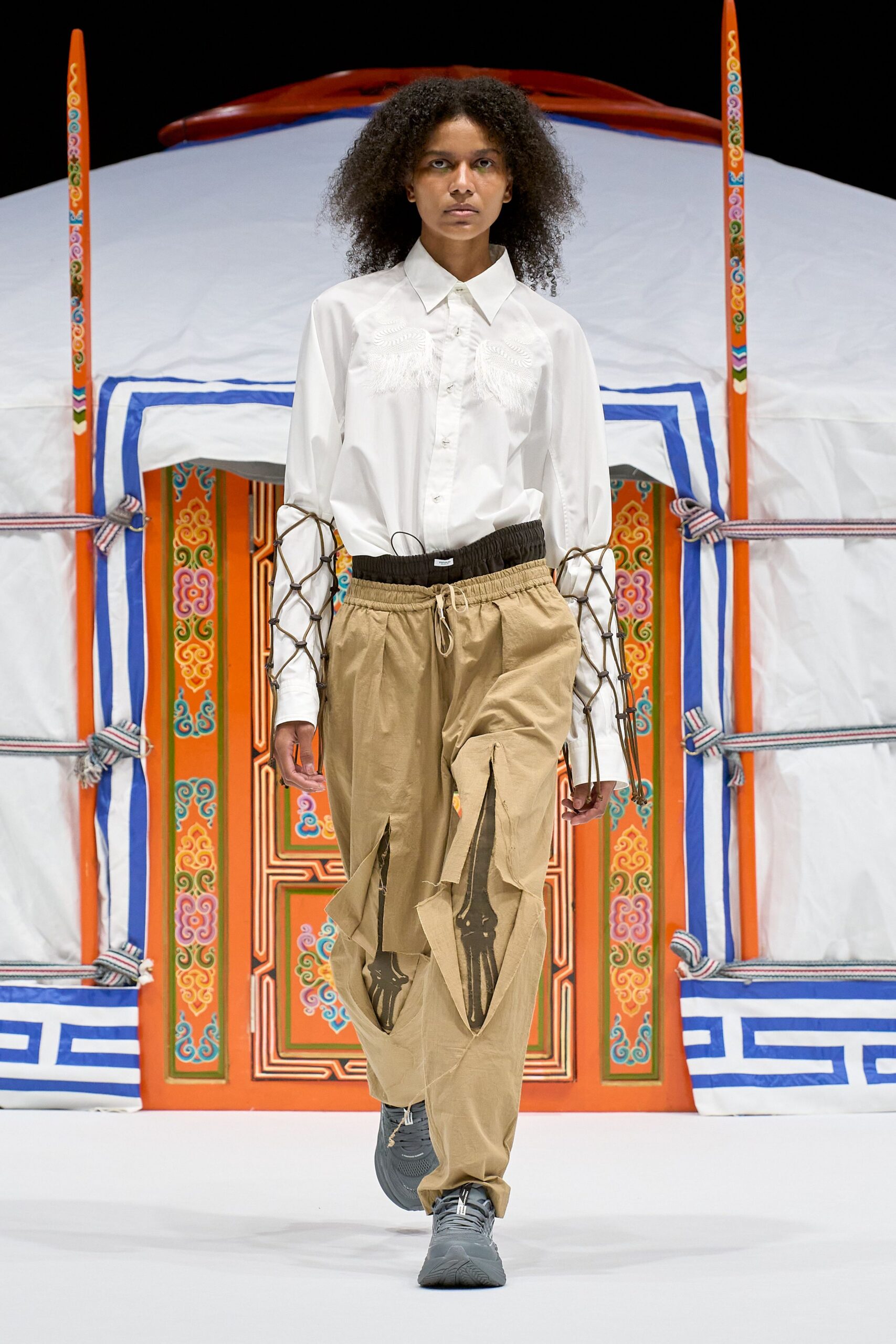 White button-up shirt with feathery appliqué at the chest and brown cord netting lashed along the sleeves. Sand drawstring trousers slashed open at the thighs over printed underlayers, gray trainers. Outside a painted yurt at noon.