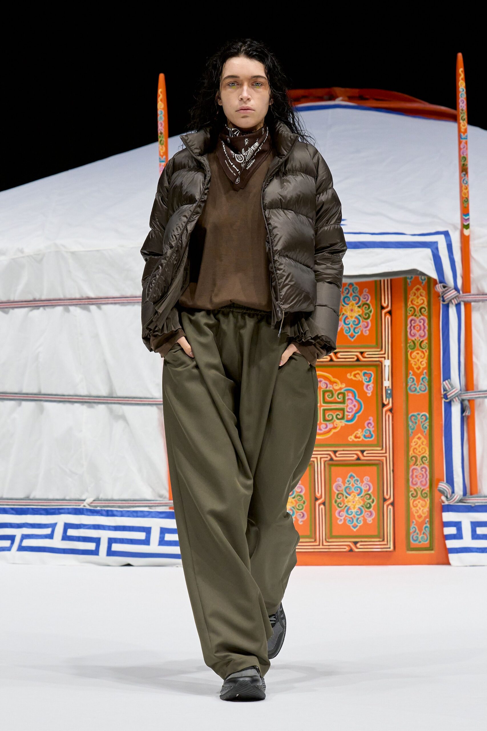 Black nylon puffer jacket with narrow horizontal baffles, dark brown jersey top, black bandana neckerchief printed in white paisley, olive draped trousers with a gathered waistband, black trainers. Dawn at a roadside yurt, air sharp with smoke.