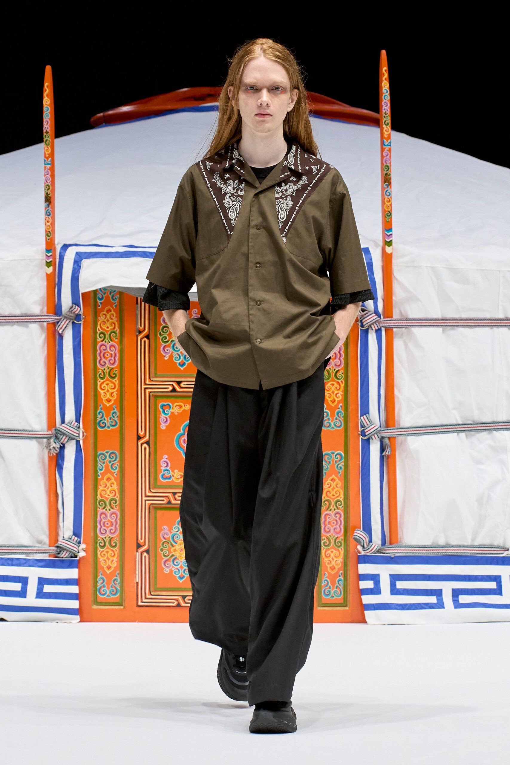 Olive camp-collar shirt with brown paisley yokes cut in sharp triangles, black mesh sleeves peeking underneath. Wide black trousers in fluid suiting, chunky black shoes. Like stepping out for air behind a lit fairground tent.