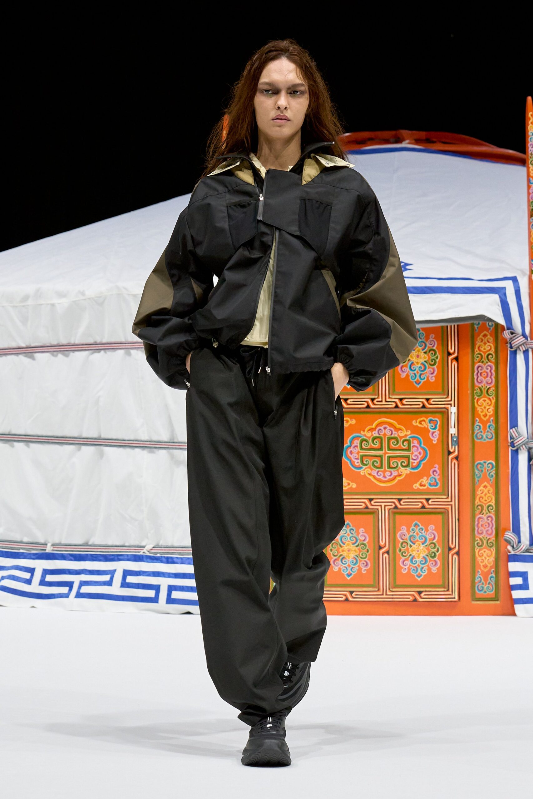 Black technical shell jacket with a folded storm flap and khaki paneling at the sleeves and hood. Loose black track pants with zip pockets and a dangling drawcord. Black trainers. Crossing a windy ferry deck before dawn.