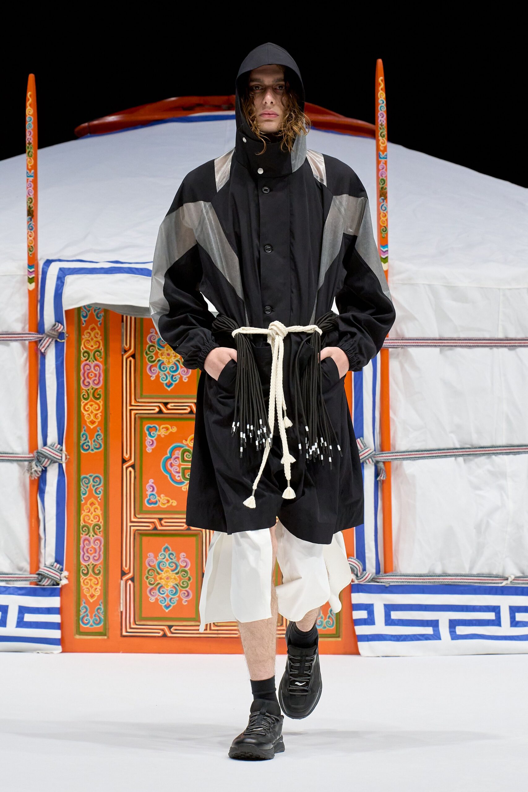 Black hooded coat with silver ripstop panels and a rope belt knotted over swinging tassel fringe tipped in white. White cropped wide-leg trousers with a fluttering split hem, black trail sneakers. Outside the yurt before dawn.