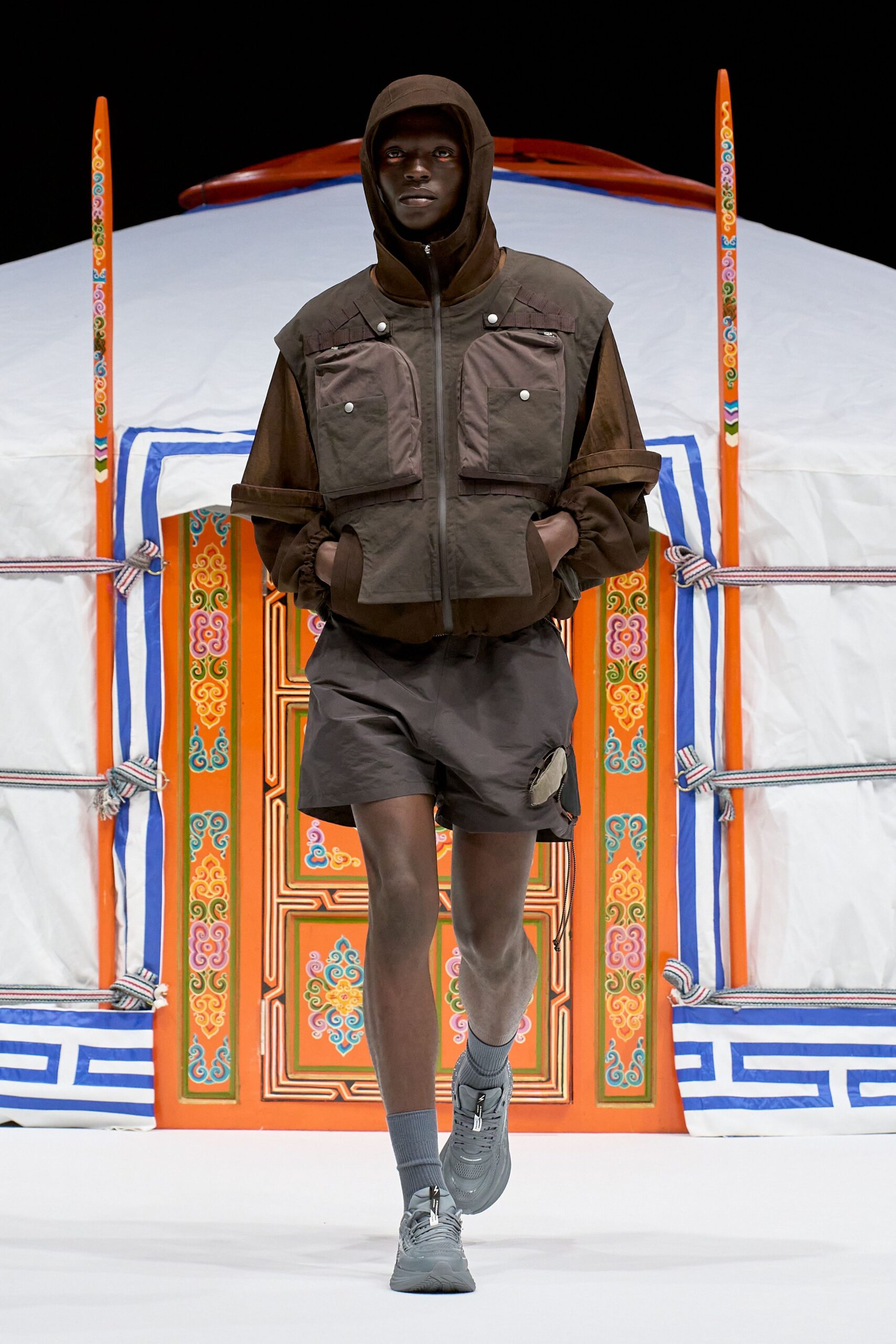 Brown hooded jacket with a zip to the chin and boxy flap pockets fixed by silver snaps, layered over rolled-sleeve top. Charcoal nylon shorts with a dangling zip pocket, grey rib socks, thick trainers. Dawn at a yurt camp, air still sharp.