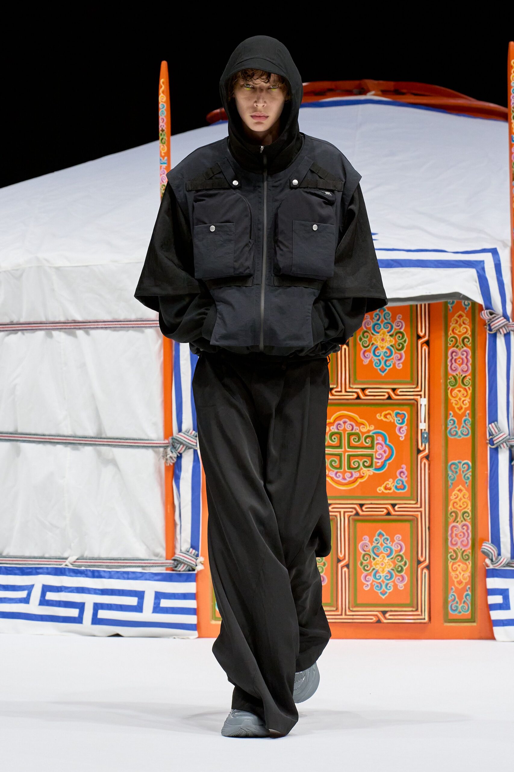 Black hooded jacket in crisp technical cloth, layered with a cropped utility vest and its silver snap pockets. Black wide-leg trousers with a deep front break, grey molded trainers. Crossing the fairground at dawn before the generators cut off.