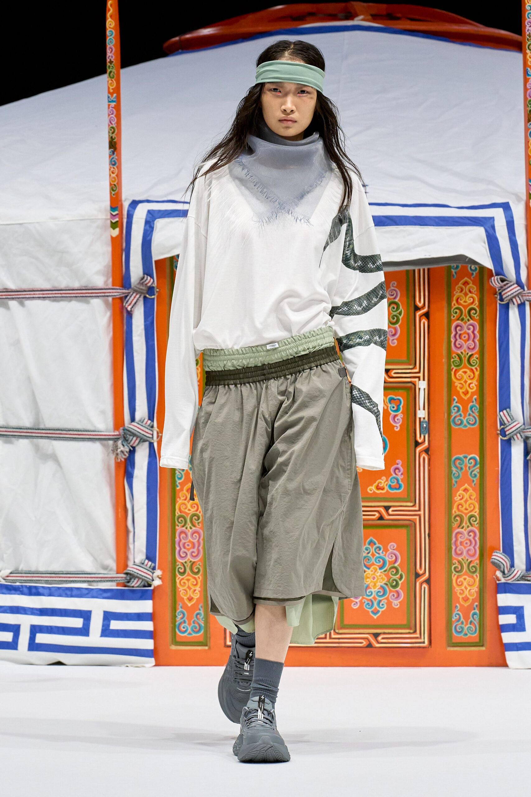 Mint jersey headband, gauzy dove-grey scarf with a fringed point, white long-sleeve top slashed with a snake-print ribbon, crinkled taupe shorts over a sheer mint underlayer, grey trainers. Wind outside the yurt flap.