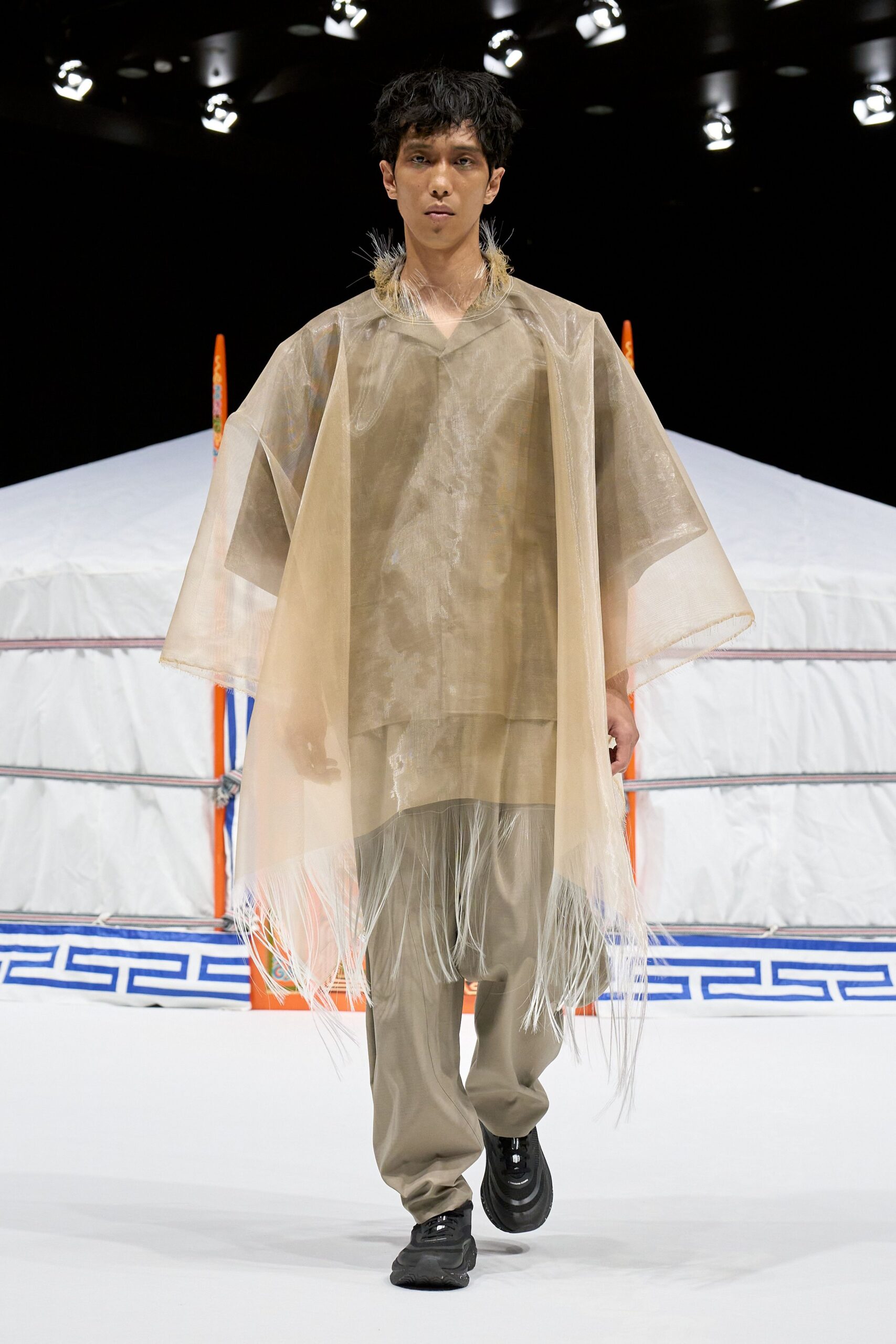 Translucent sand organza poncho with fine fringe shaking at the hem, over a matte taupe V-neck tunic and loose trousers with a soft break at the ankle, finished with black rubber trainers. Like stepping out of a steam tent at dawn.