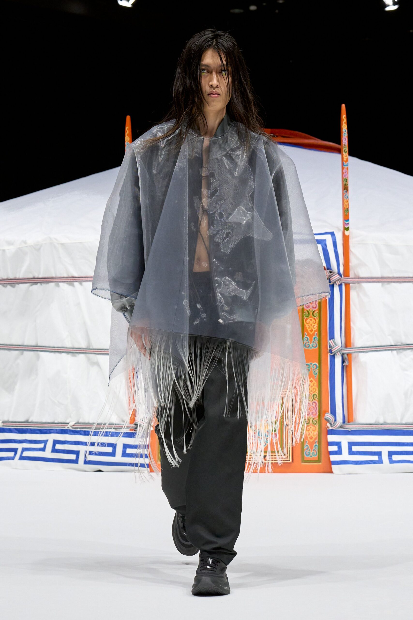Smoky sheer poncho in stiff organza, its hem dropping into white horsehair fringe over a black glossy jacket. Bare chest with a fine cord tie, black straight-leg trousers, black trainers. Outside a tent at dawn, grass still wet.