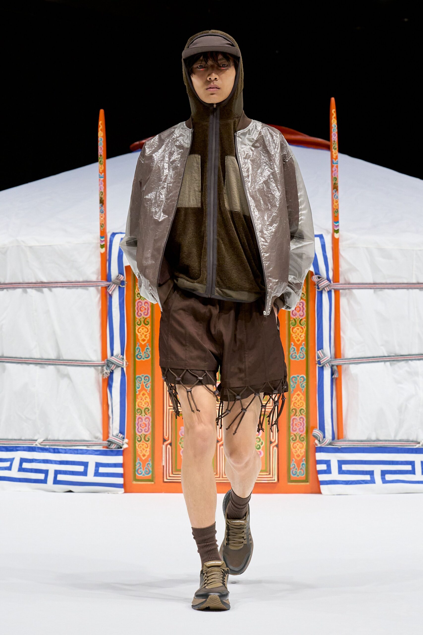 Taupe visor, dark olive fleece zip hoodie with pale chest panels, silver crinkled shell bomber, and espresso shorts finished with dangling bungee lattice at the hem. Ribbed socks, trail sneakers. Dawn outside a wind-battered camp.