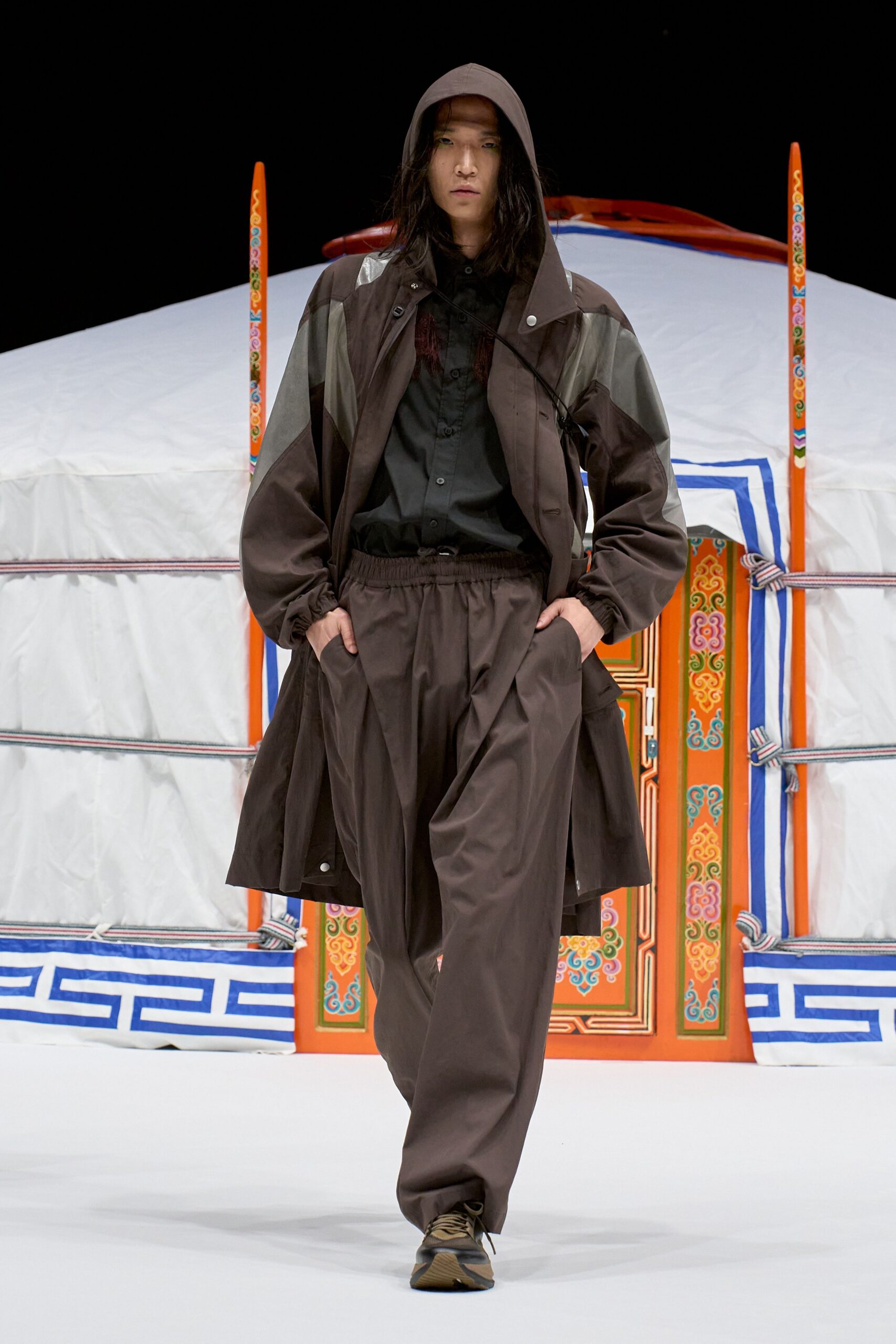 Hooded brown shell coat with pale grey paneling and snap-tab lapel, over a black button shirt. Billowy drawstring trousers in the same matte shell, trail-running sneakers. Like stepping out before dawn at a roadside yurt.