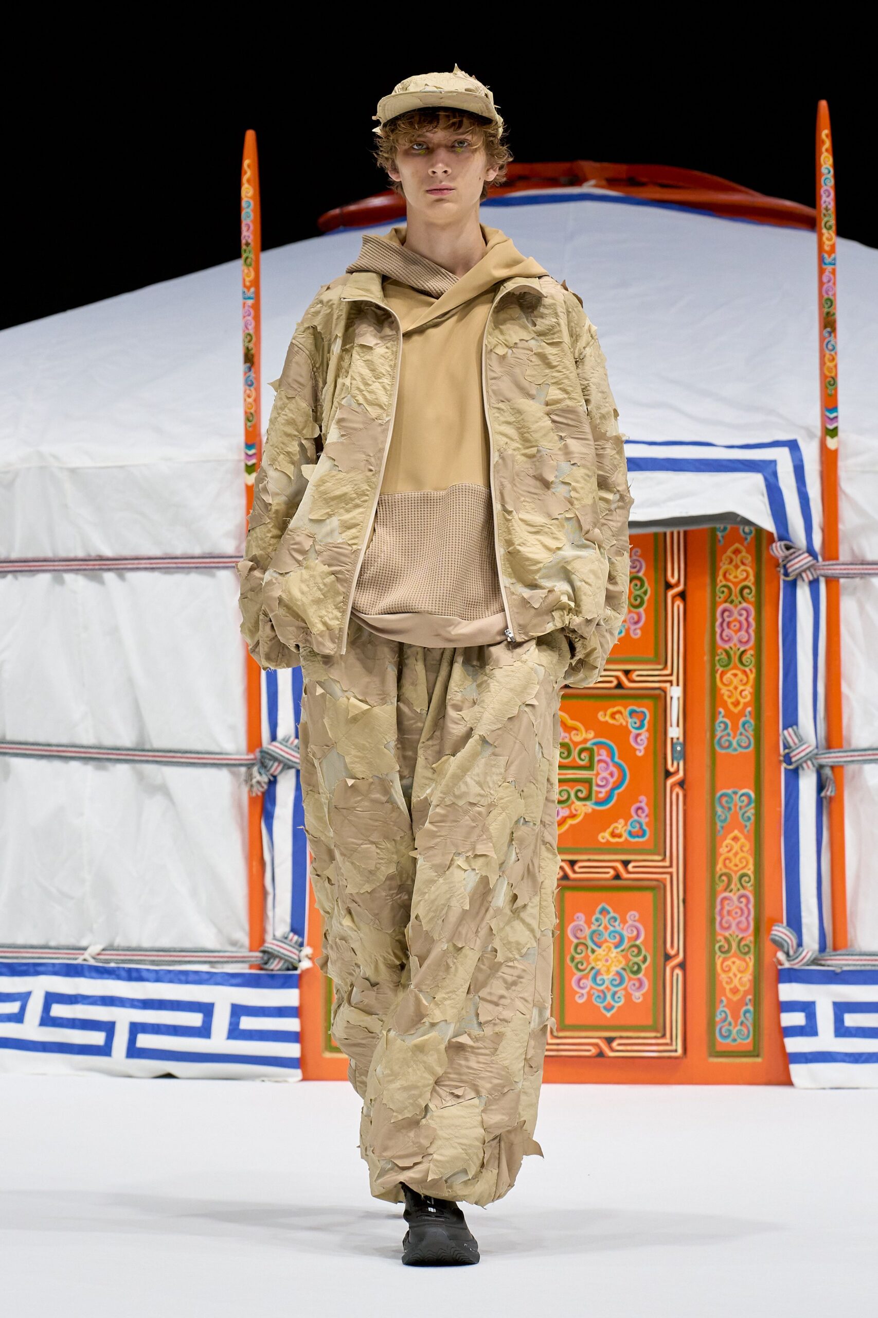 Sand cap with a frayed brim, beige hooded top with a waffle-knit pouch, crinkled zip jacket layered in peeling appliqued strips, matching loose trousers, black trainers. Like stepping out of a yurt at dawn.