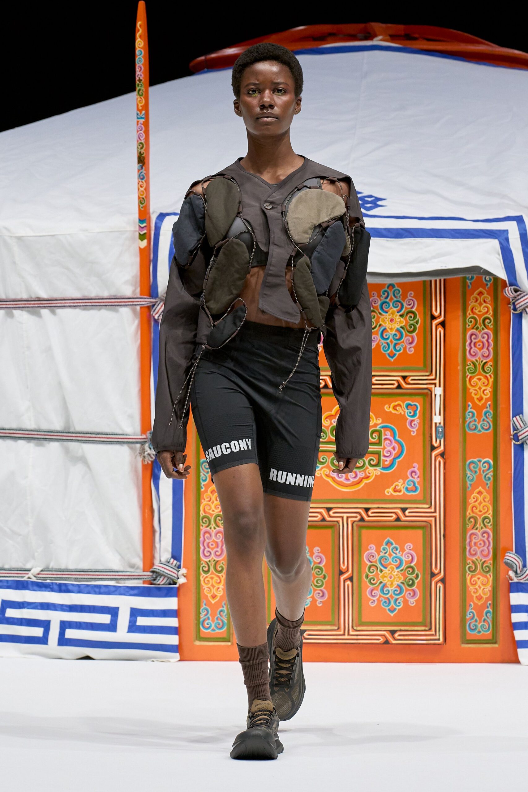 Cropped smoke-grey shirt jacket with padded oval patches linked by exposed zip tape and a single off-center button. Black compression cycling shorts with rubberized lettering, ribbed socks, trail runners. Dawn miles past the last campsite.