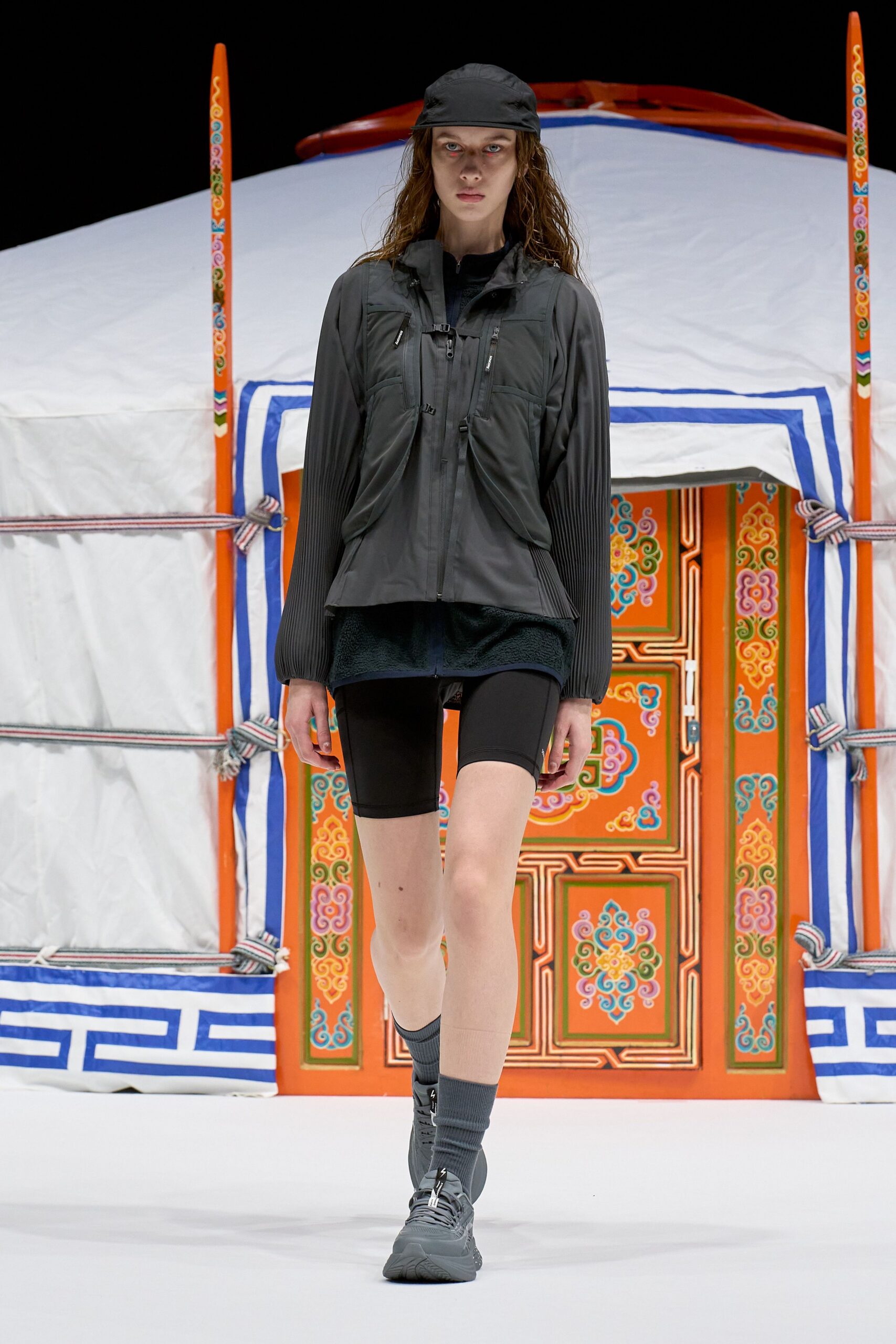 Black nylon cap, charcoal shell jacket with pleated sleeves and twin zip chest pockets clipped by a sternum strap, black fleece tunic, matte cycling shorts, ribbed socks, grey trail sneakers. Dawn at the yurt camp before the stove catches.