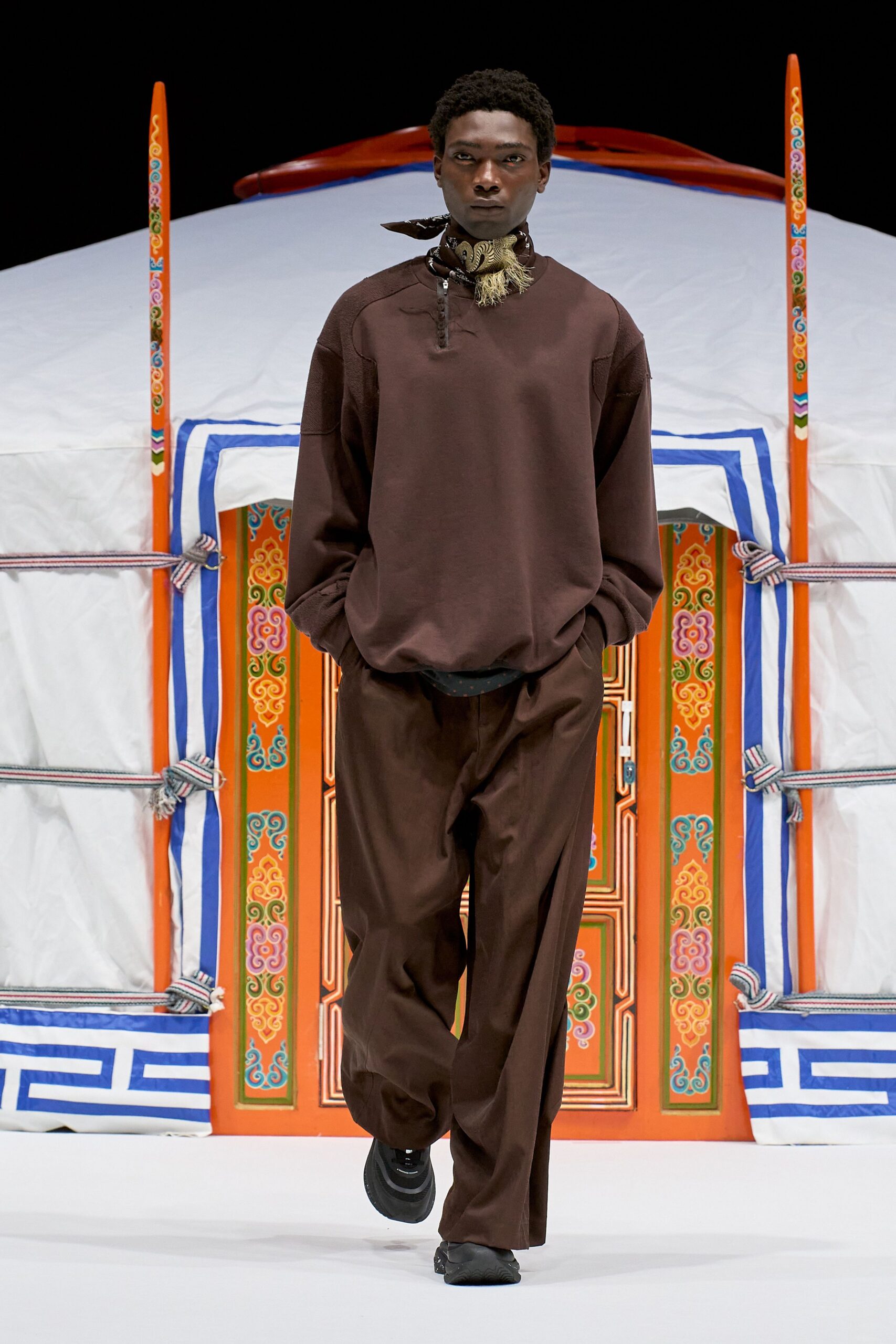 Brown jersey pullover with a short half-placket and dropped seams, silk neck scarf knotted with a tasseled end. Loose brown trousers with a soft break, black trainers. Cold morning outside a painted yurt, hands still warm in pockets.