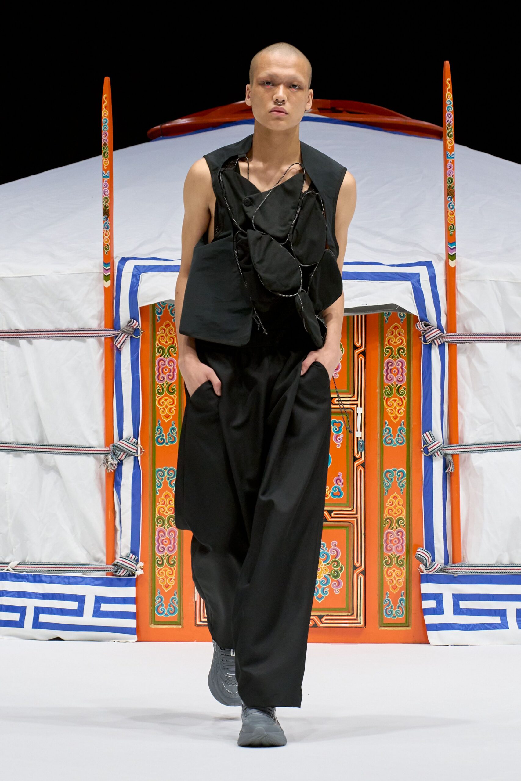 Black sleeveless top built from overlapping pouch-like panels, each edged with cord piping and one loose tab hanging at the hip. Wide black trousers with deep slant pockets, charcoal knit trainers. Outside the tent at dawn, hands still cold.