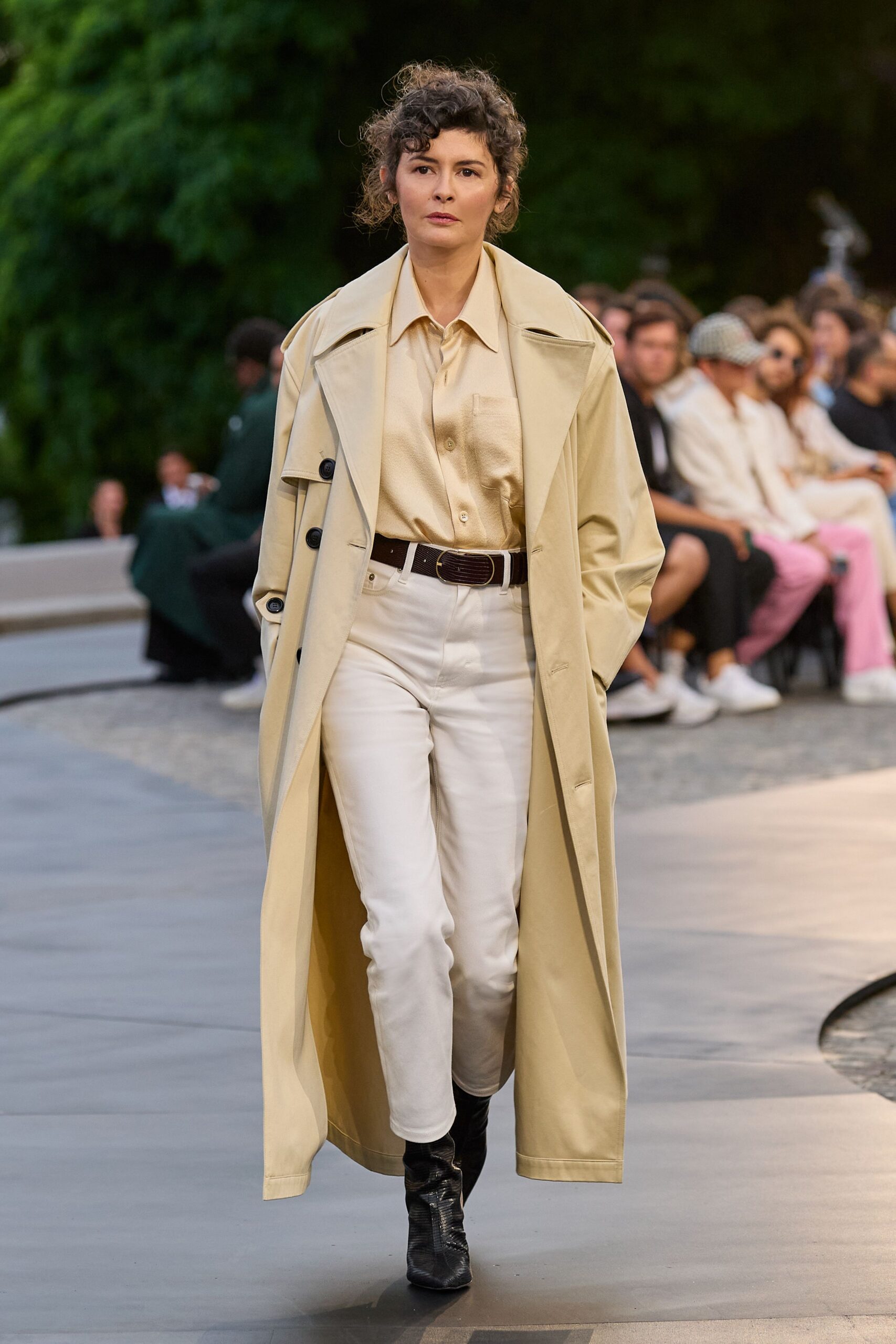 Buttercream trench coat with broad storm flaps and black cuff buttons. Champagne satin shirt, one chest pocket catching the light. Dark brown leather belt with an oval buckle. Ecru straight jeans, cropped just above the ankle. Black croc-embossed ankle boots. Garden runway at dusk, when the air still holds the day's heat.