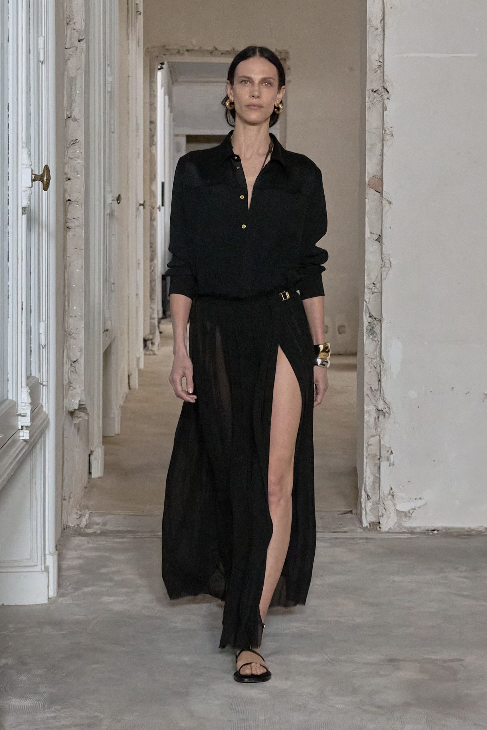Thick gold hoops hugging both lobes. Black shirt in fluid crepe, gold buttons stopping below the sternum. Sheer black wrap skirt, pleats falling from a side-buckled waist. Hammered gold cuff at one wrist. Flat black thong sandals. Crossing a half-renovated apartment before the dinner guests arrive.