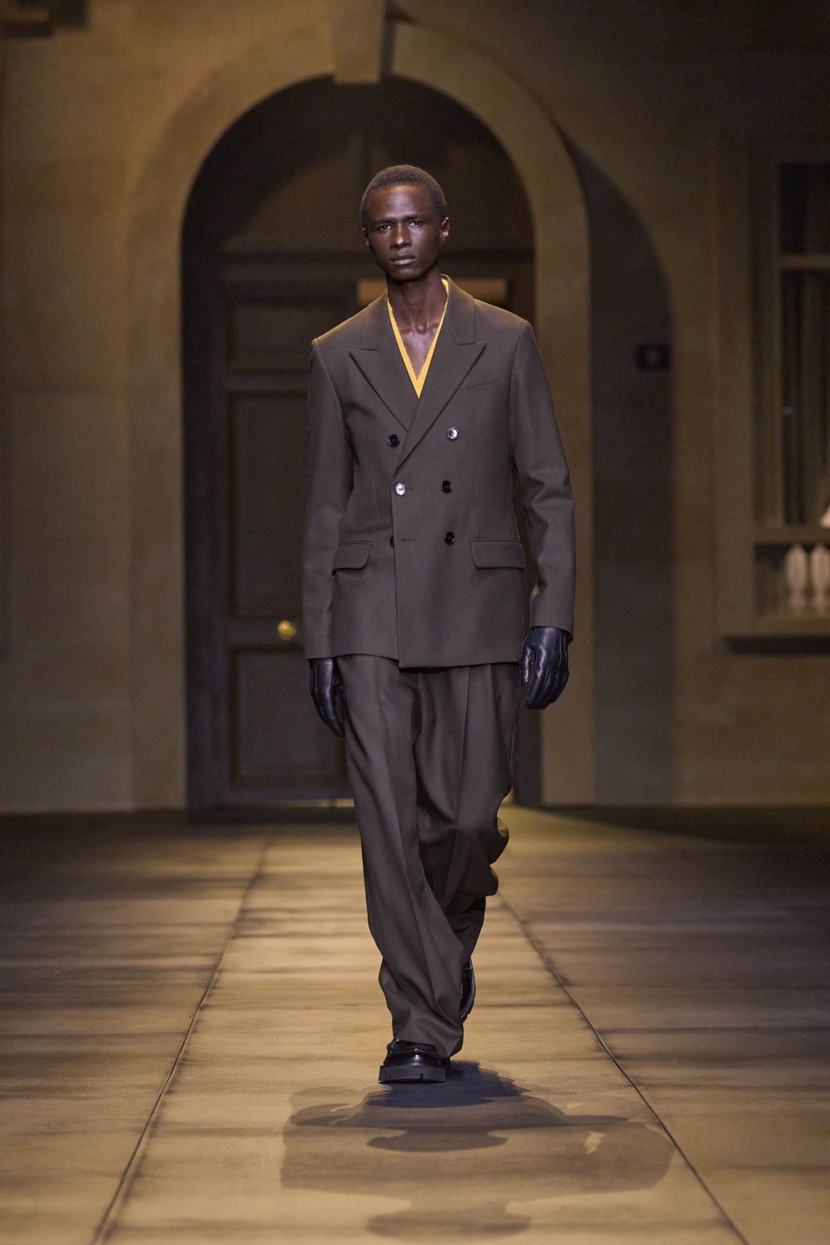 Espresso double-breasted blazer with broad peaks and one pale horn button. Marigold V-neck layer cut narrow at the chest. Matching wool trousers falling loose from a soft front pleat. Black leather gloves. Square-toe black shoes. For crossing a stone courtyard before dinner is called.