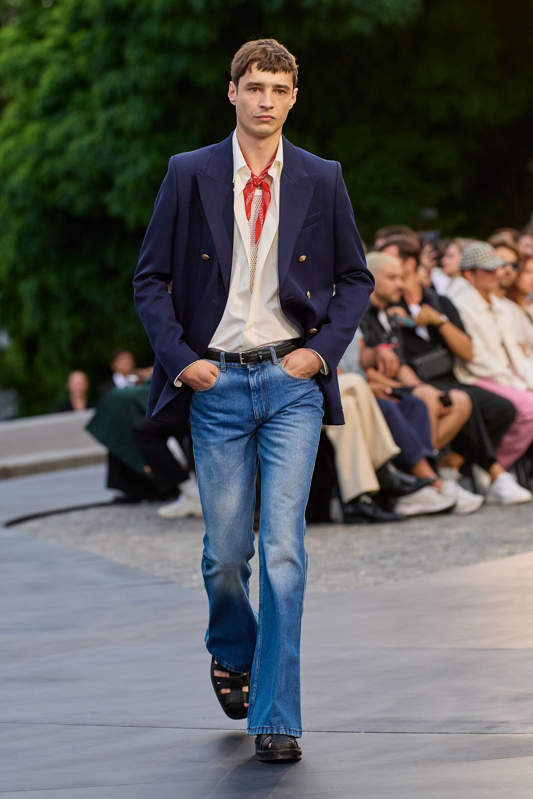 Navy double-breasted blazer with broad peak lapels and gilt buttons. Cream shirt worn open over a narrow strip of mesh. Red paisley neckerchief knotted close under the collar. Light-wash bootcut jeans, whiskered pale through the thighs. Black leather fisherman sandals. Walking across a hotel terrace just before dinner service.