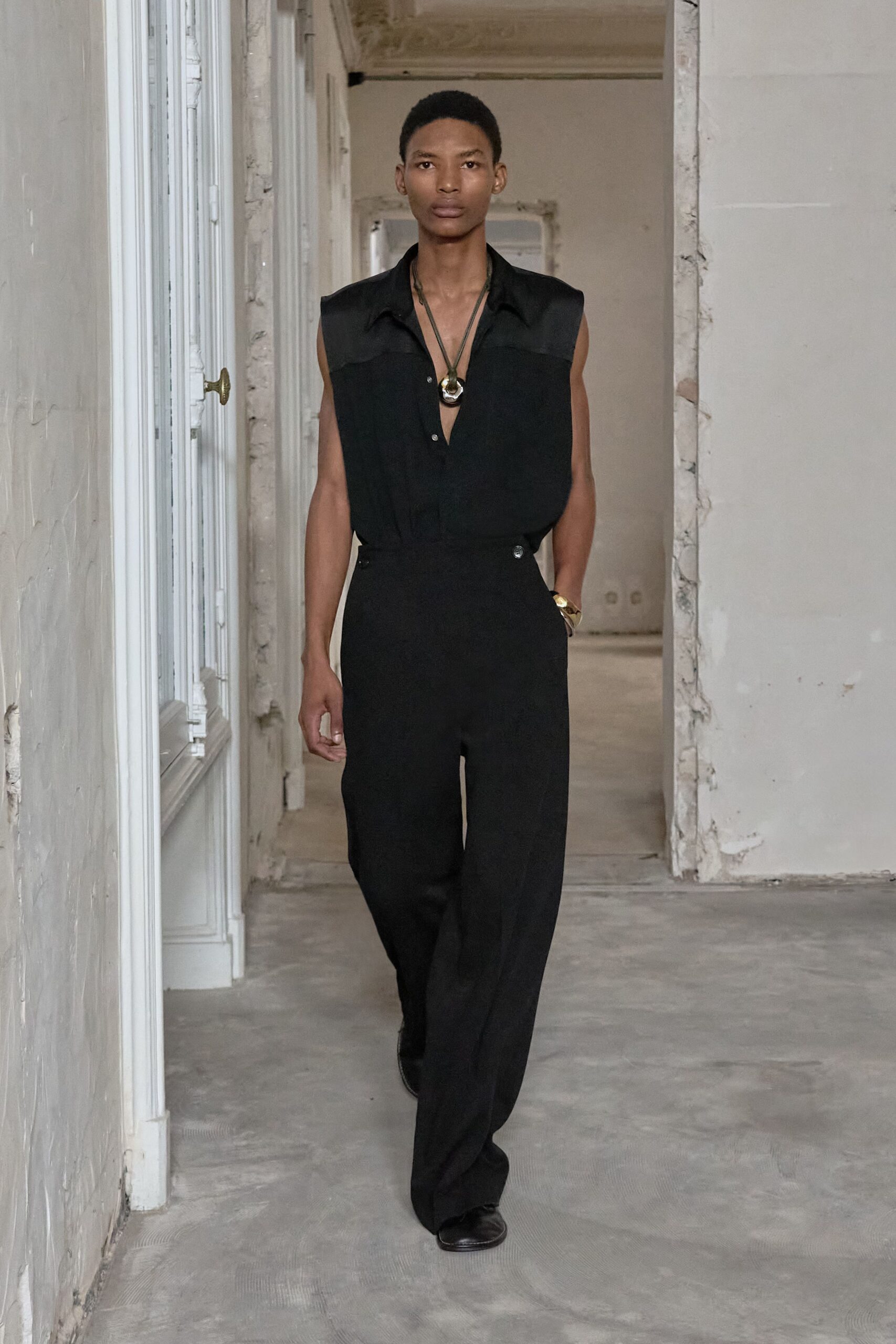 Black fluid sleeveless shirt with a deep placket and soft collar. Long cord necklace with a chunky silver-and-gold pendant. High-waisted black trousers, side tabs buttoned tight at the hip. Wide gold cuff bracelet. Black leather shoes, squared off at the toe. Walking through an empty apartment before the plaster dust settles.