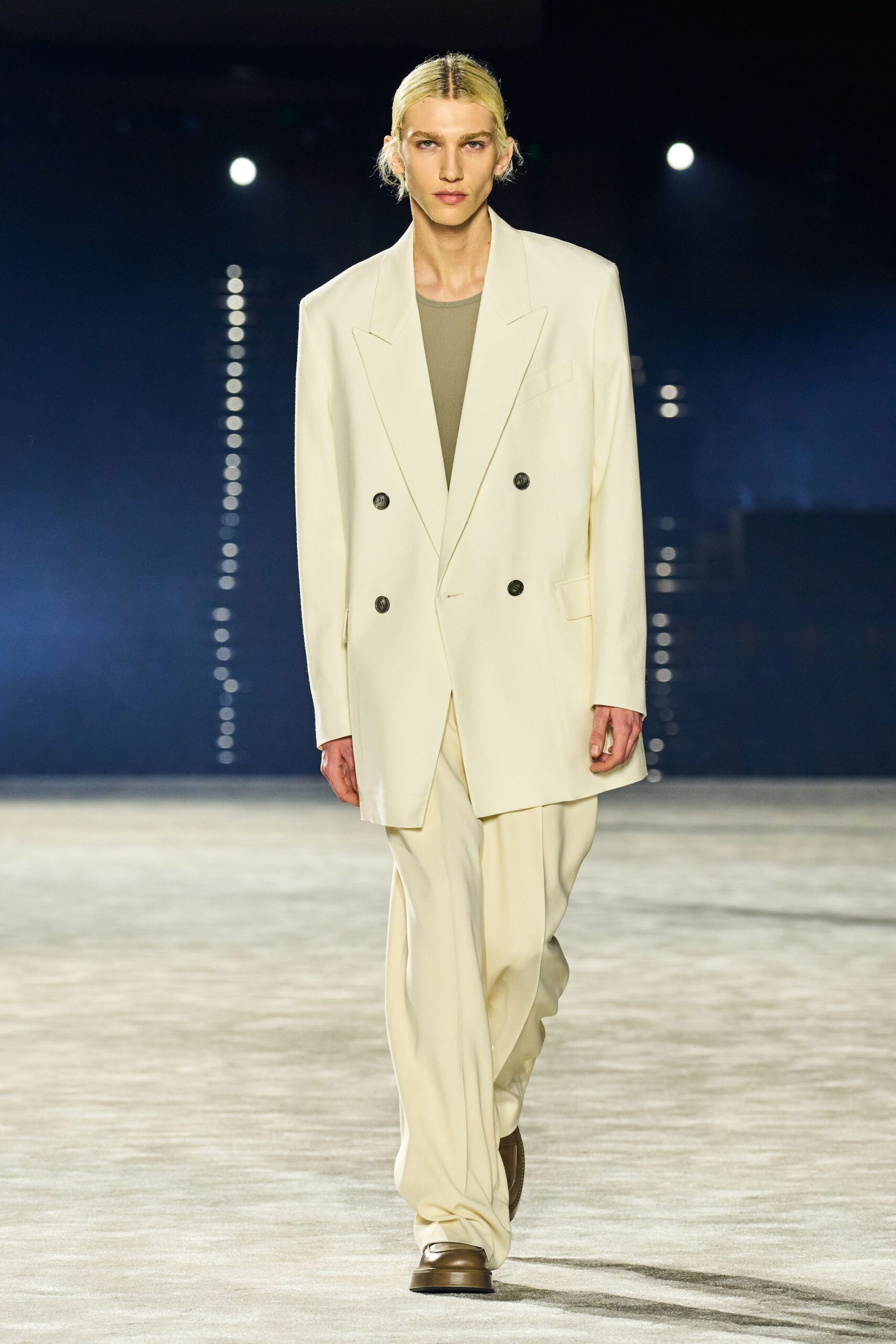 Long ivory double-breasted blazer with broad peak lapels and dark horn buttons. Moss ribbed tank close at the throat. Fluid ivory trousers breaking into a soft puddle over the shoe. Square-toe brown leather shoes with a thick raised welt. Early checkout at a lakeside hotel, suit still creased from the car.