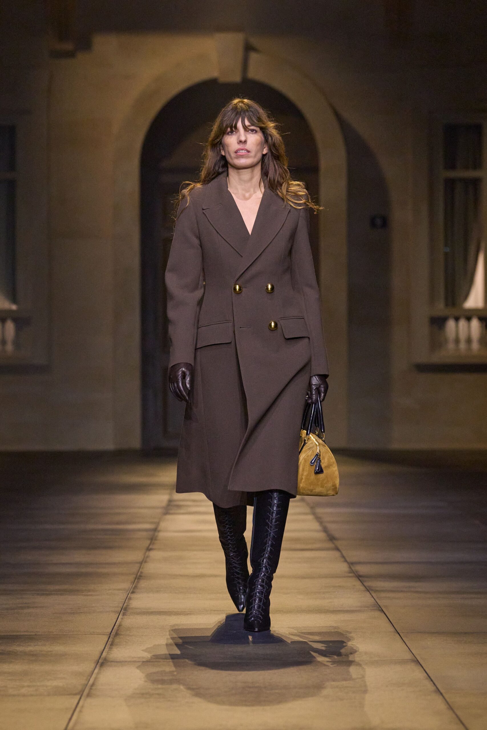 Chocolate wool double-breasted coat, one brass button sitting oddly low. Dark plum leather gloves. Mustard suede handbag with glossy black rolled handles. Black knee-high boots, tightly ladder-laced up the front. Crossing a stone courtyard after dusk to return somebody else's keys.
