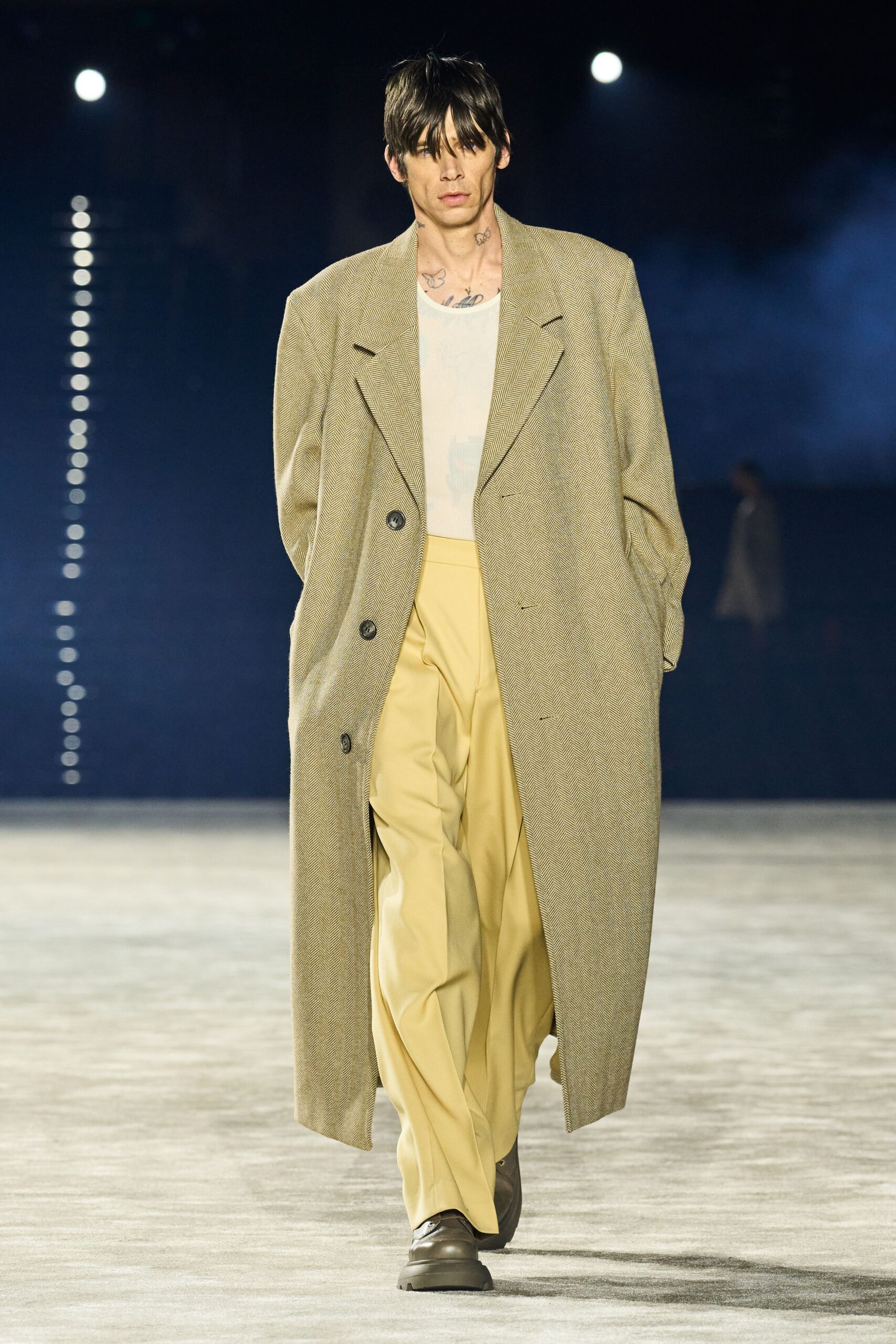 Long taupe herringbone overcoat with broad peak lapels and slumped shoulders. Sheer ivory ribbed tank, tattoos ghosting through the chest. Butter yellow tailored trousers falling in a soft break. Mud-brown leather shoes with thick rounded soles. Whatever you pull on for a dawn train after not going home.