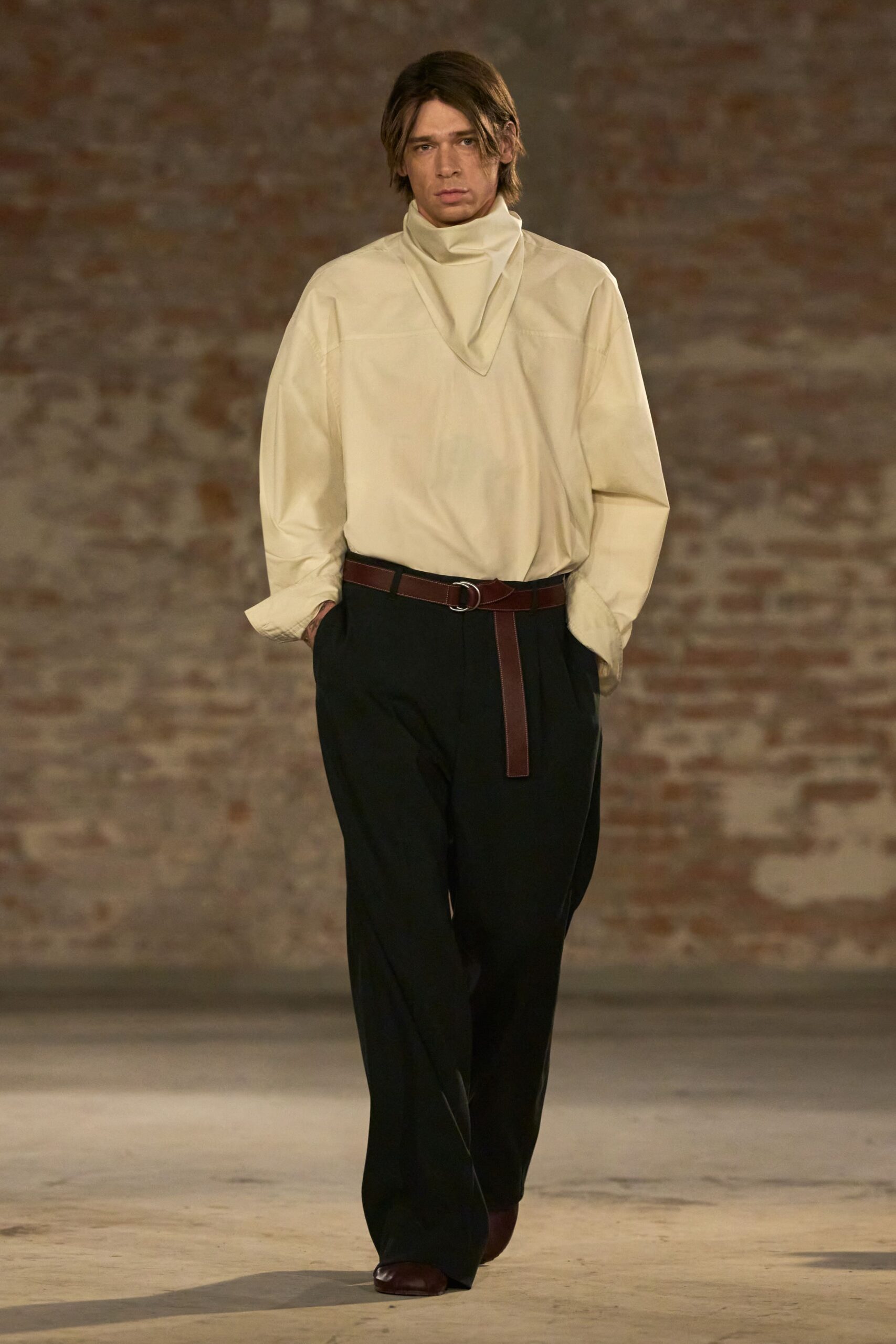 Cream tunic in crisp poplin, built with a folded funnel scarf collar. Oxblood leather belt with a silver ring buckle and long hanging tongue. Black wide-leg trousers breaking softly over the shoe. Oxblood square-toe shoes. Walking across a drafty hall after dinner service has ended.