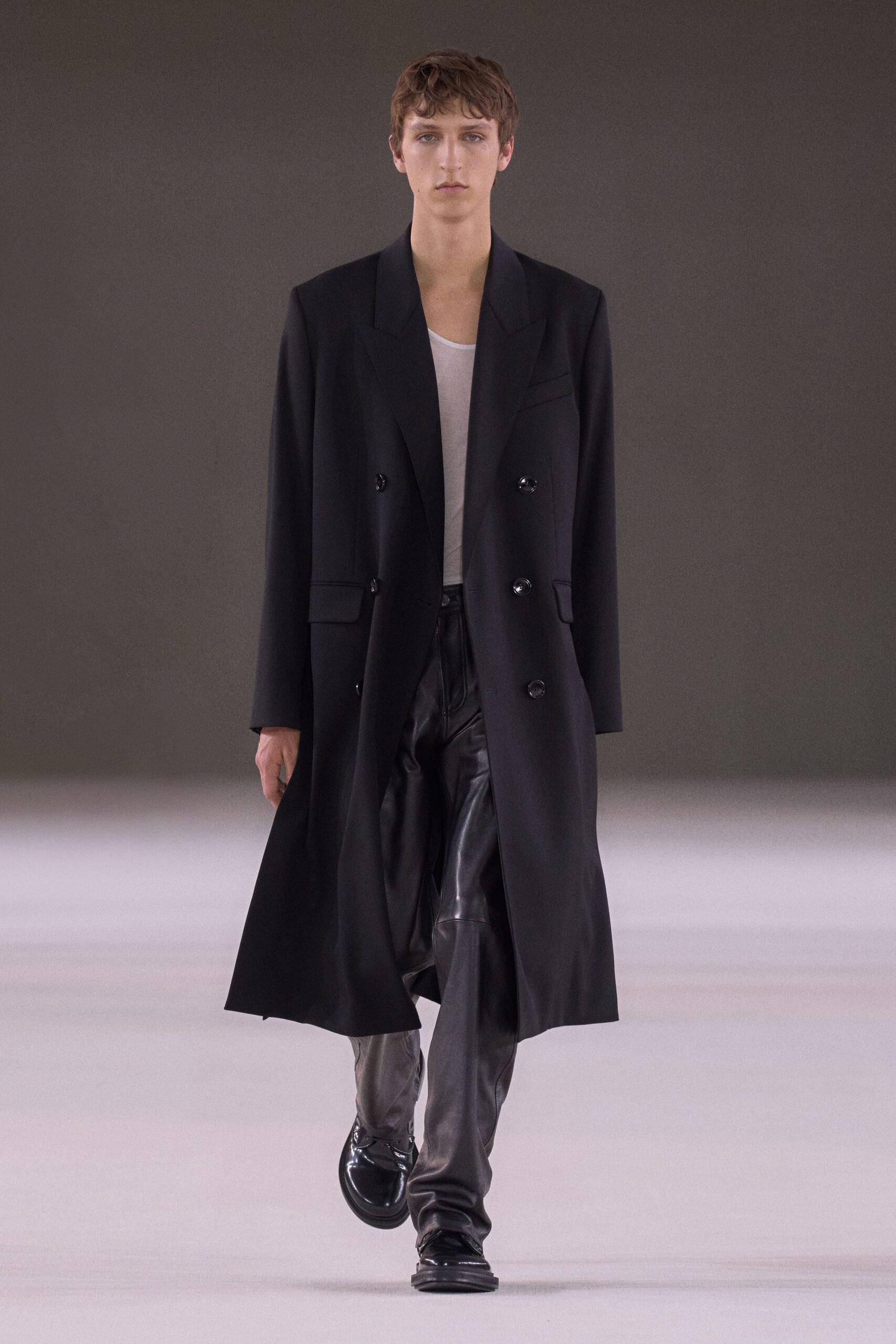 Black double-breasted coat with broad peaked lapels and glossy buttons. White scoop-neck tank in thin semi-sheer jersey. Black leather trousers falling in long creases over the shoes. Black rounded loafers with a mirror-bright finish. Crossing an empty hotel lobby before sunrise checkout.