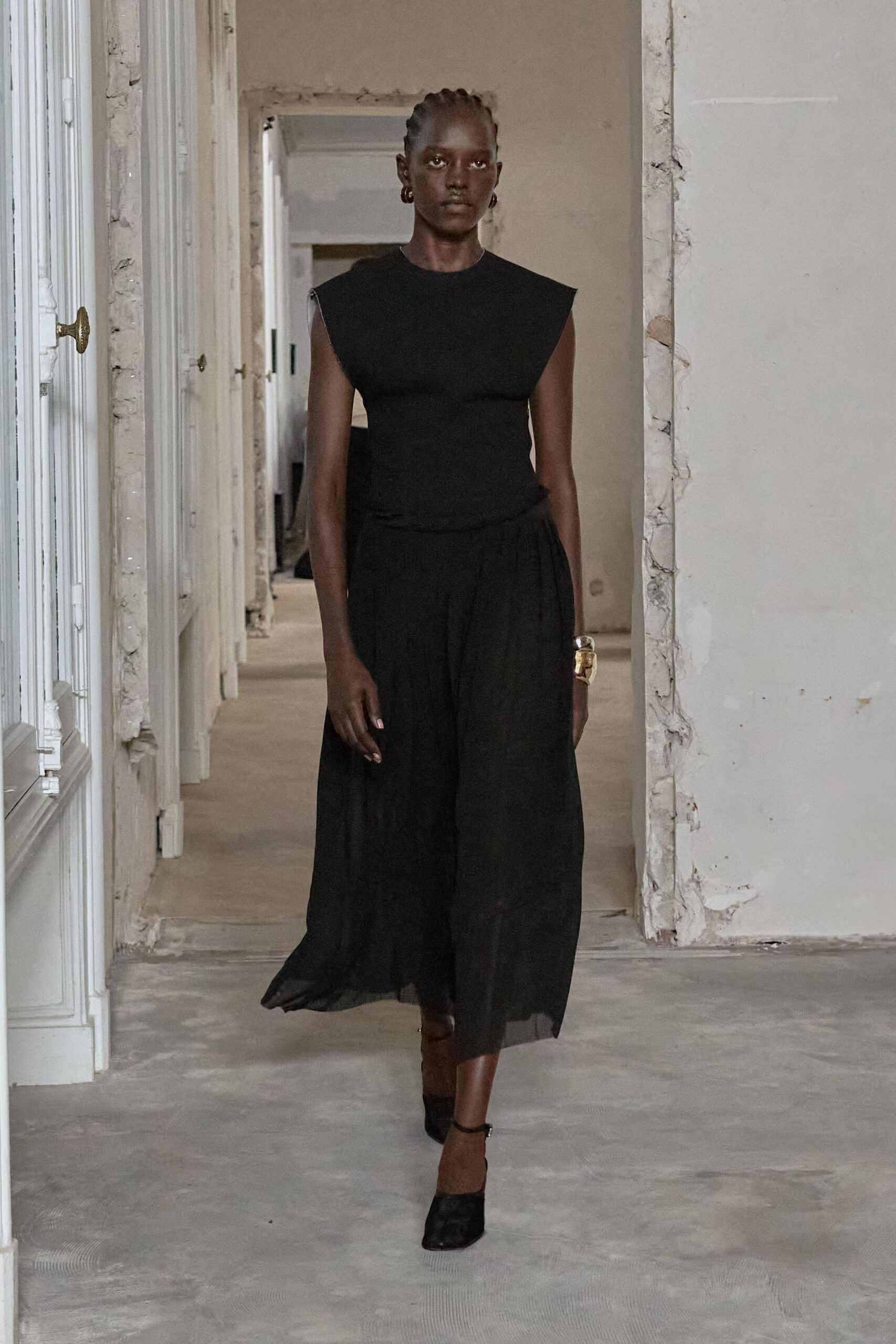 Black knit shell with squared cap sleeves jutting past the shoulders. Ink gauze midi skirt, softly gathered and going sheer at the hem. Wide hammered gold cuff. Black ankle-strap heels. Crossing a half-renovated apartment before the dust sheets come off.
